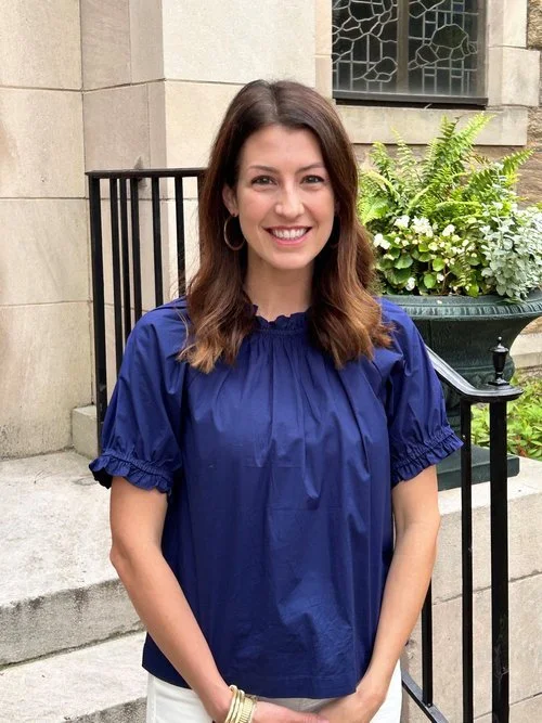 A woman with brown hair wearing a blue blouse and white skirt, smiling, standing outdoors in front of a stone building with a black railing and a large potted plant.
