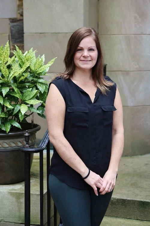 A woman with shoulder-length brown hair, wearing a black sleeveless blouse and dark pants, standing outdoors next to a potted plant with green and yellow leaves.
