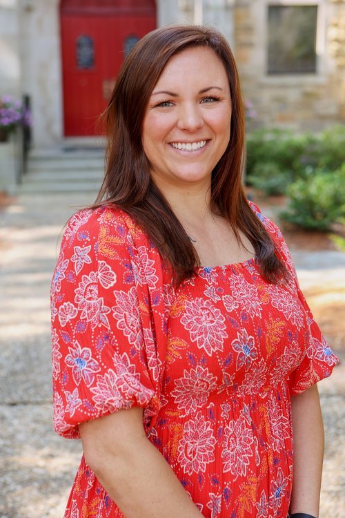 A woman with long brown hair smiling outdoors in front of a stone building with a red door, wearing a red floral dress.