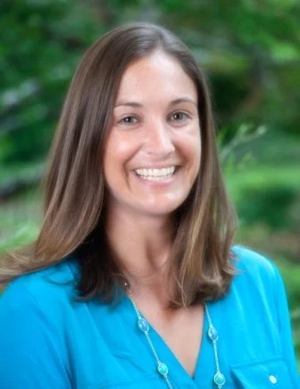 A woman with long brown hair smiling, wearing a blue top and a necklace, outdoors with green foliage in the background.