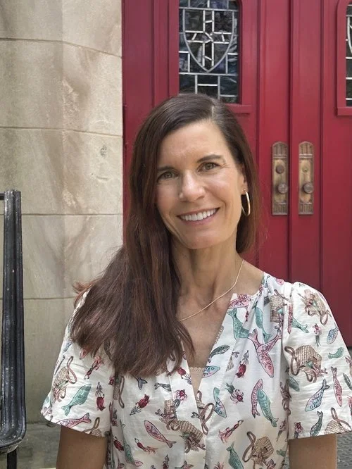 Woman with long brown hair and hoop earrings smiling in front of red doors and beige stone wall.