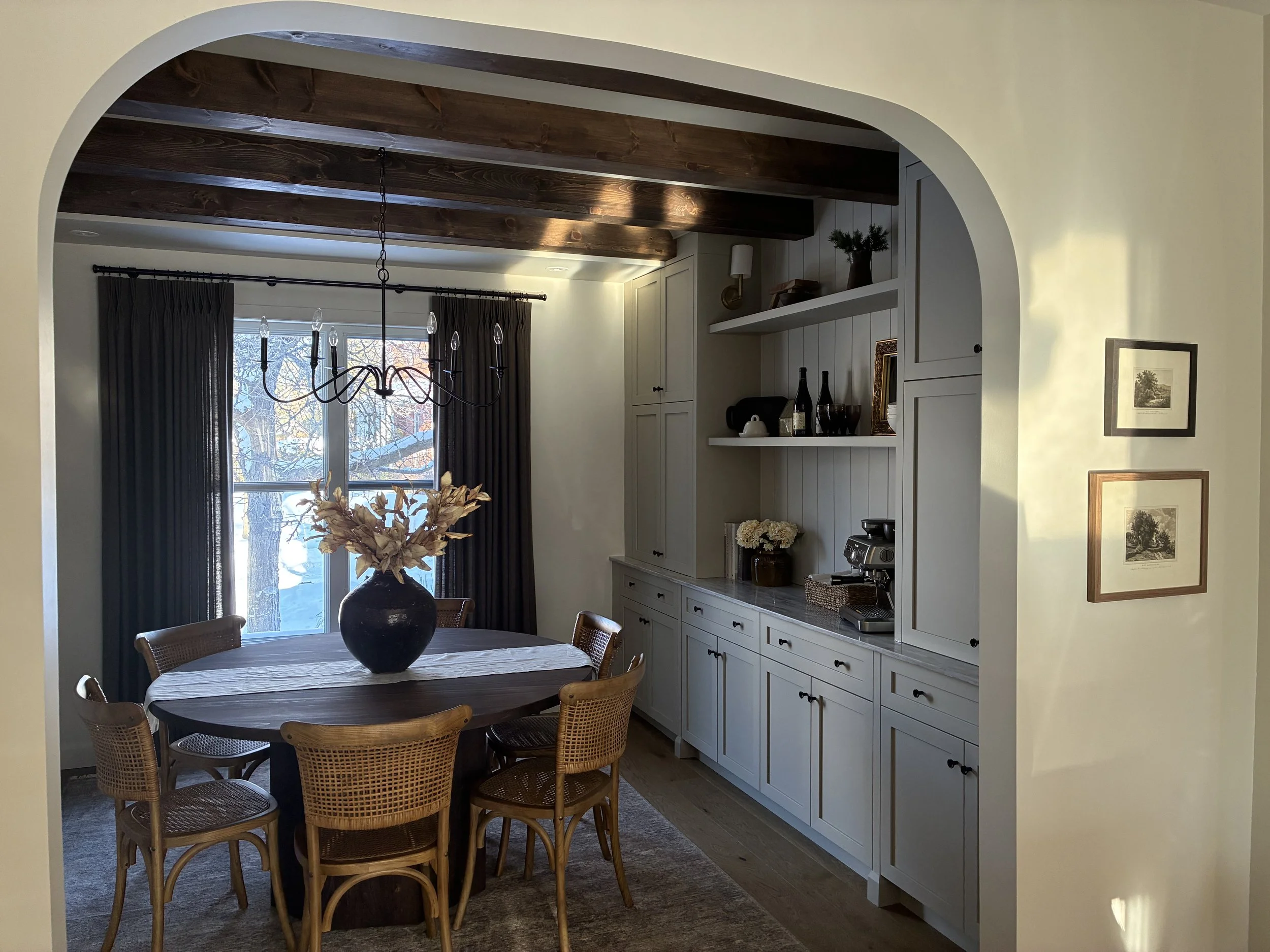 Salle à manger avec une table ronde en bois, entourée de six chaises en rotin, et un vase de fleurs séchées sur la table. En arrière-plan, une grande fenêtre avec des rideaux noirs, un chandelier noir suspendu au plafond, et un placard aménagé en ray