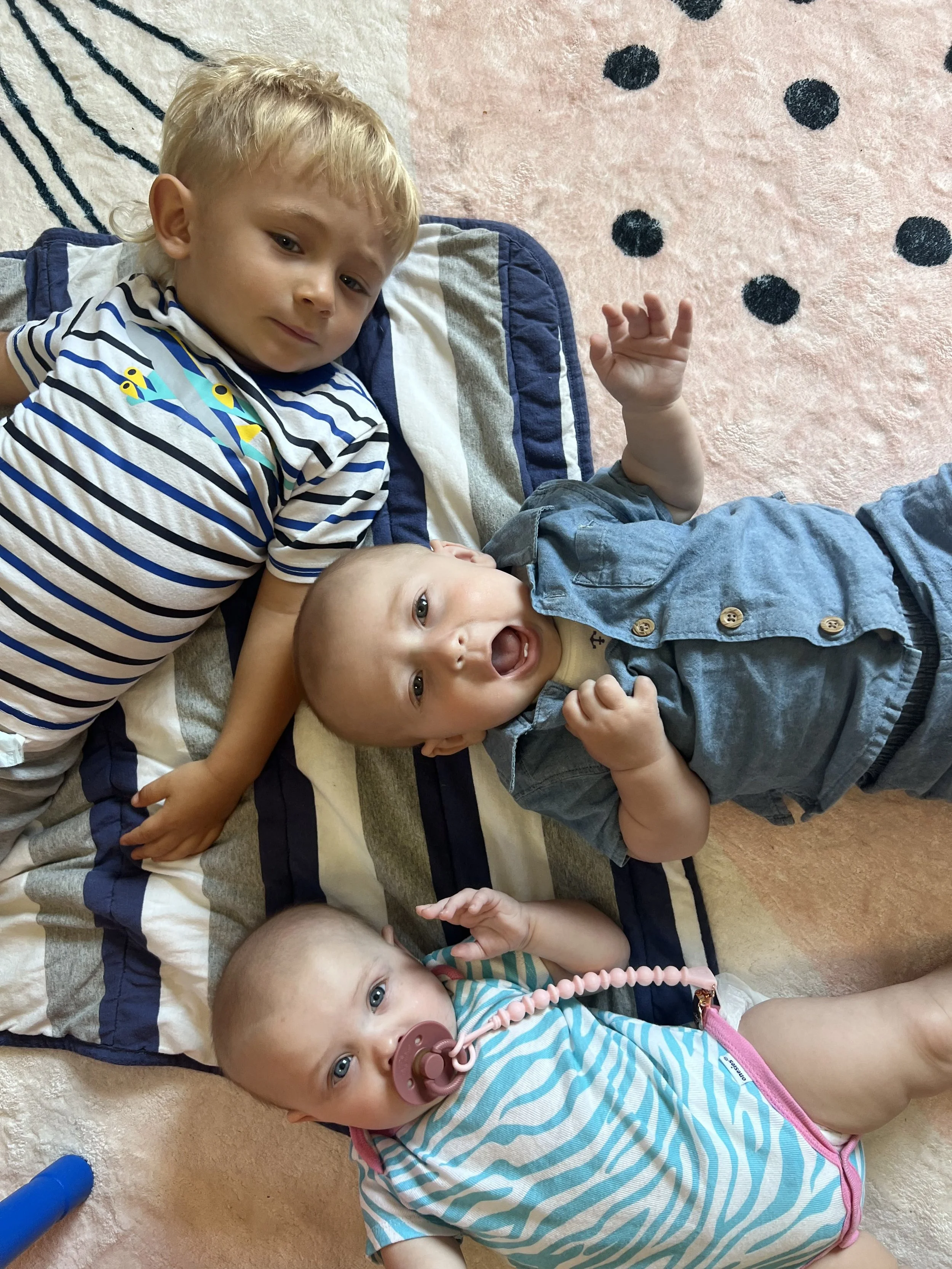 Three young children lying on a striped blanket on the floor. The oldest child is in a striped shirt, the middle child has a blue shirt and is smiling, and the youngest baby has a pacifier and a striped onesie.