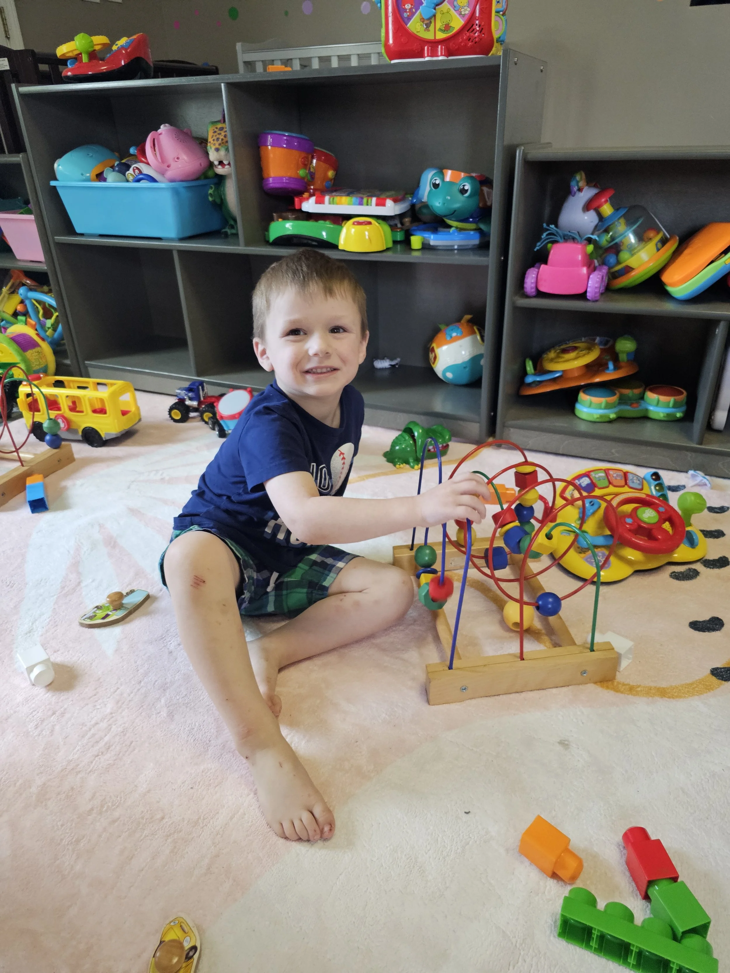 A young boy sitting on the floor of a playroom, playing with a bead maze toy, surrounded by various colorful toys and storage shelves filled with more toys.