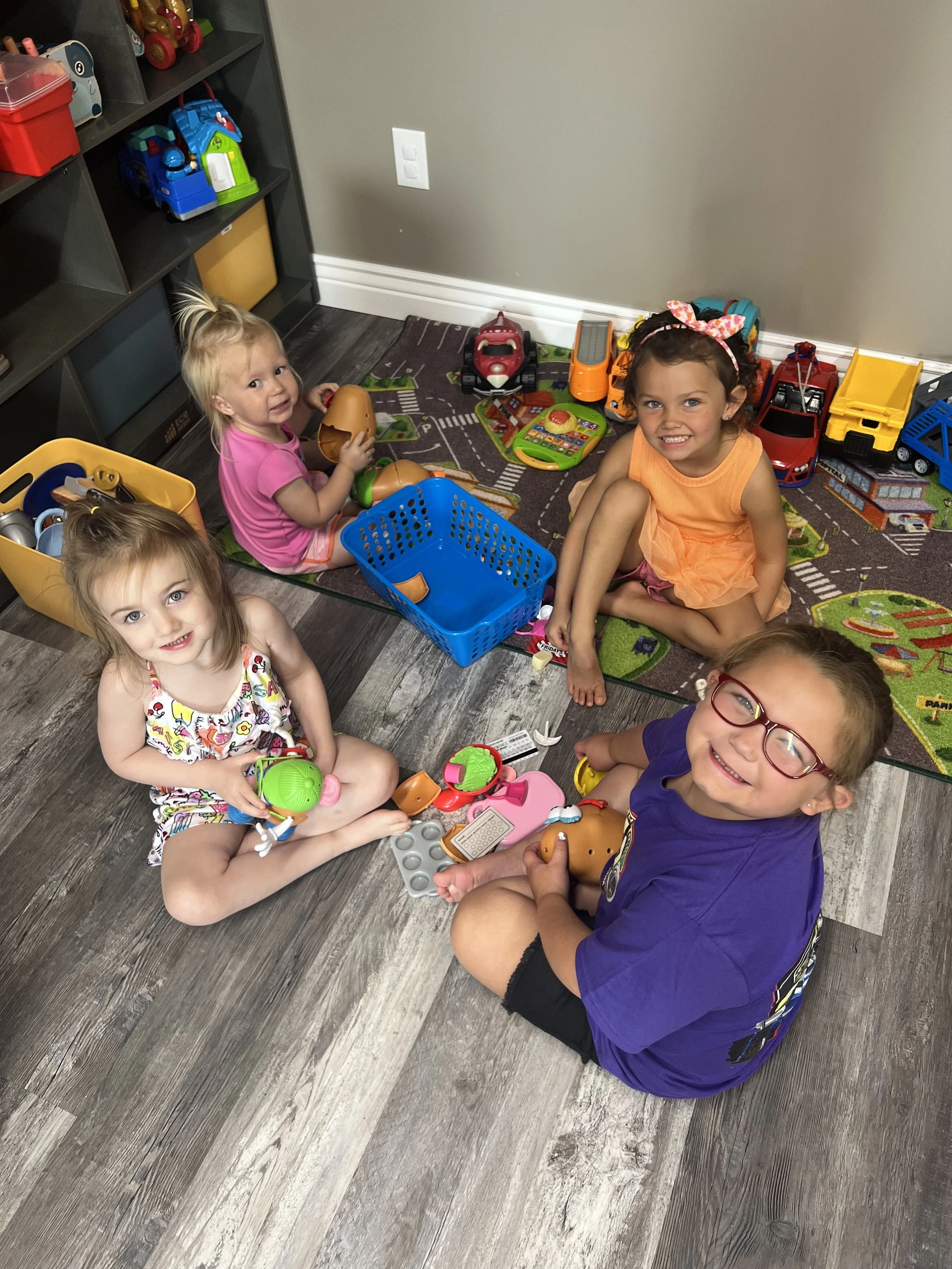 Four children sitting on a floor surrounded by toy toys with a play area and shelves behind them.