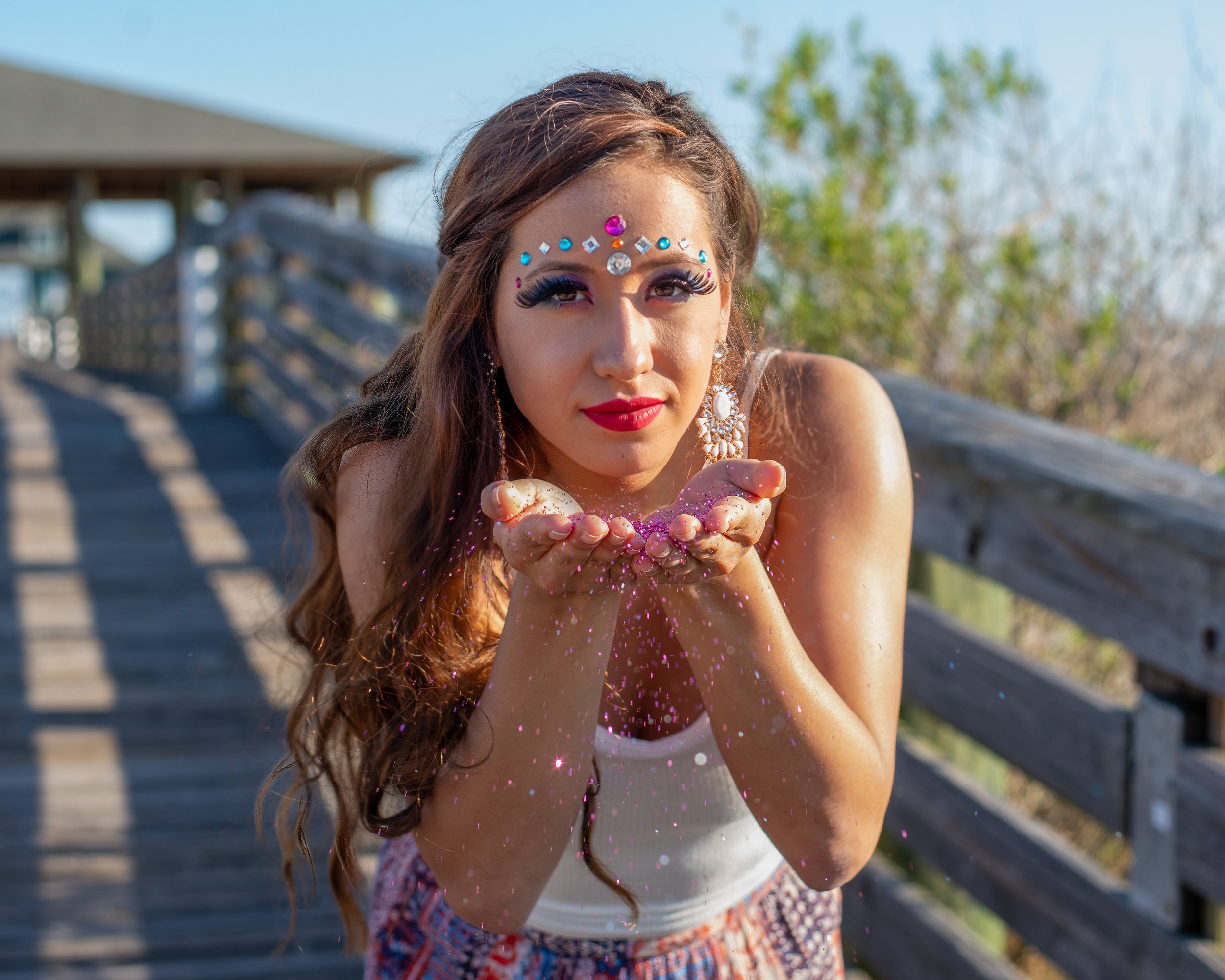 Young woman with long hair decorated with colorful rhinestones and makeup, blowing pink glitter from her hands outdoors.
