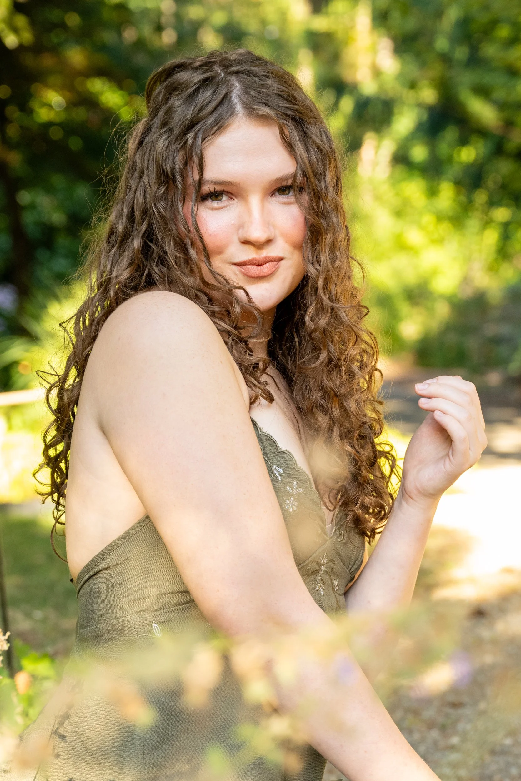 A senior portrait of woman with curly brown hair and light skin is outdoors in a wooded area, looking at the camera with a slight smile. She is wearing a khaki-colored dress with floral embroidery and has one arm bent with her hand near her shoulder.