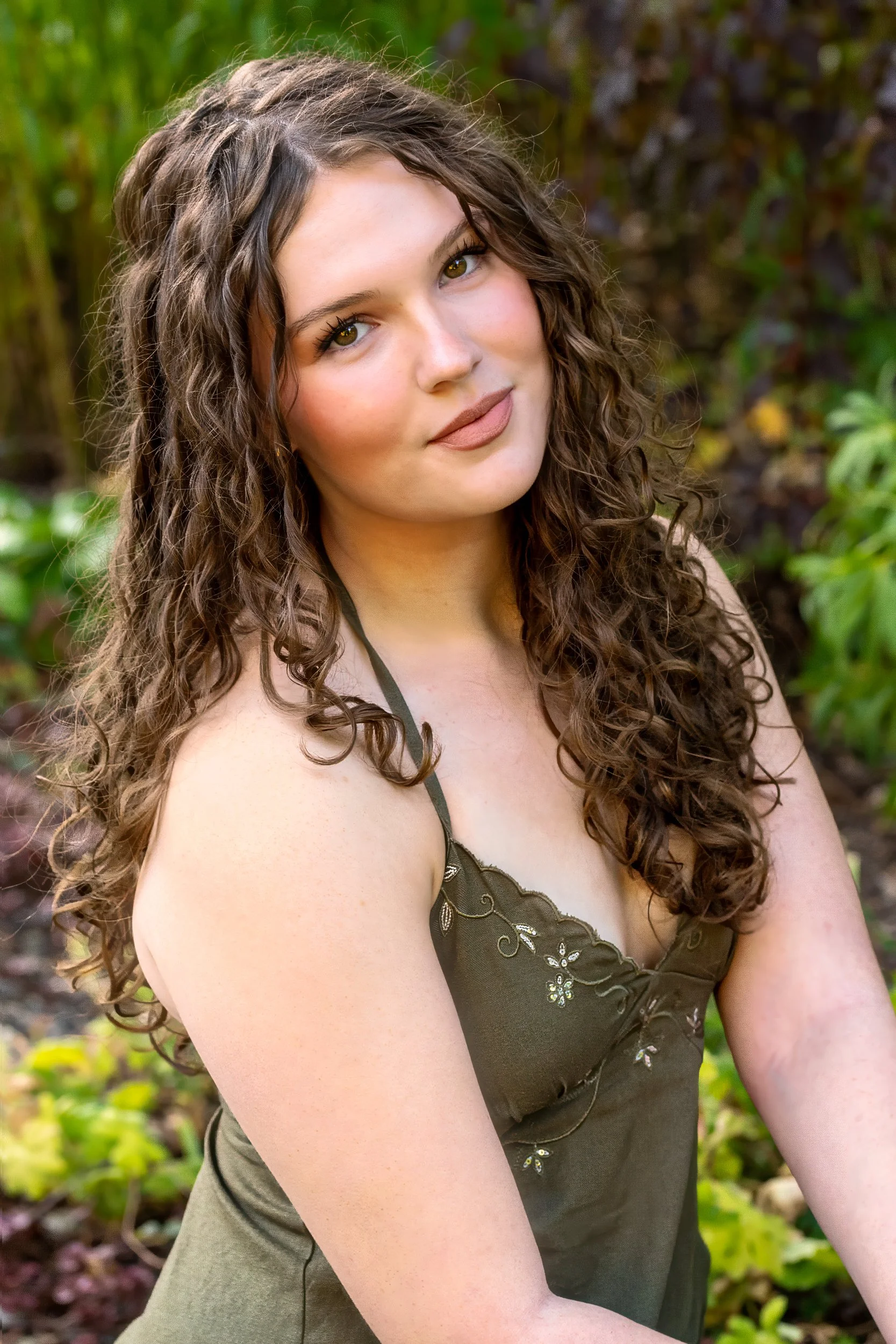 A senior portrait of a young woman with long curly brown hair and light skin, wearing a dark green dress with embroidered floral details, posing outdoors with a background of green foliage.