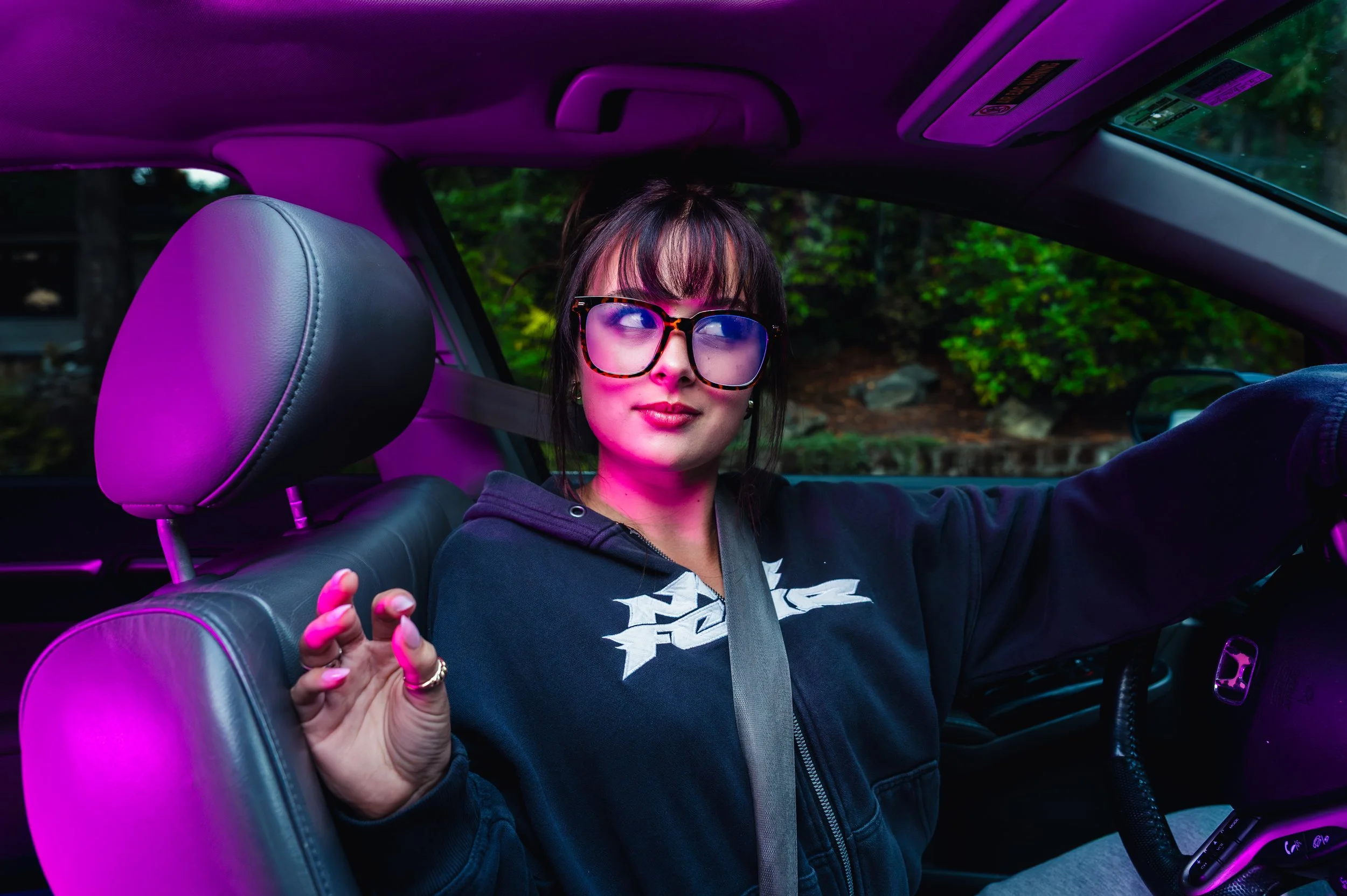 Young woman with dark hair wearing large tortoiseshell glasses, black hoodie, and seatbelt, sitting in the driver seat of a car with purple interior lighting, with green foliage visible through the window.