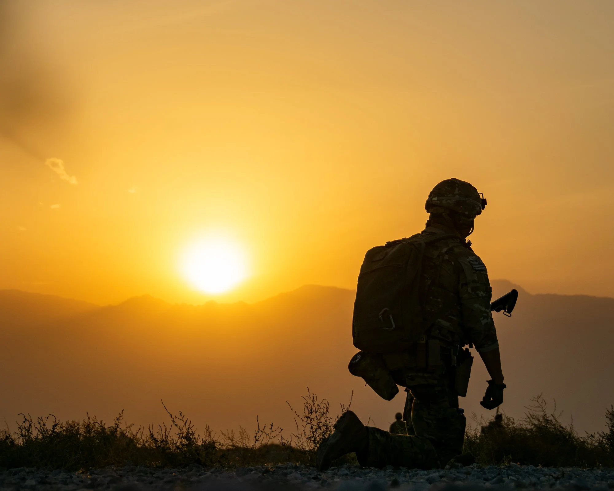 Soldier in camouflage uniform standing outdoors during sunset, holding a firearm with a scenic mountain and sunset background.
