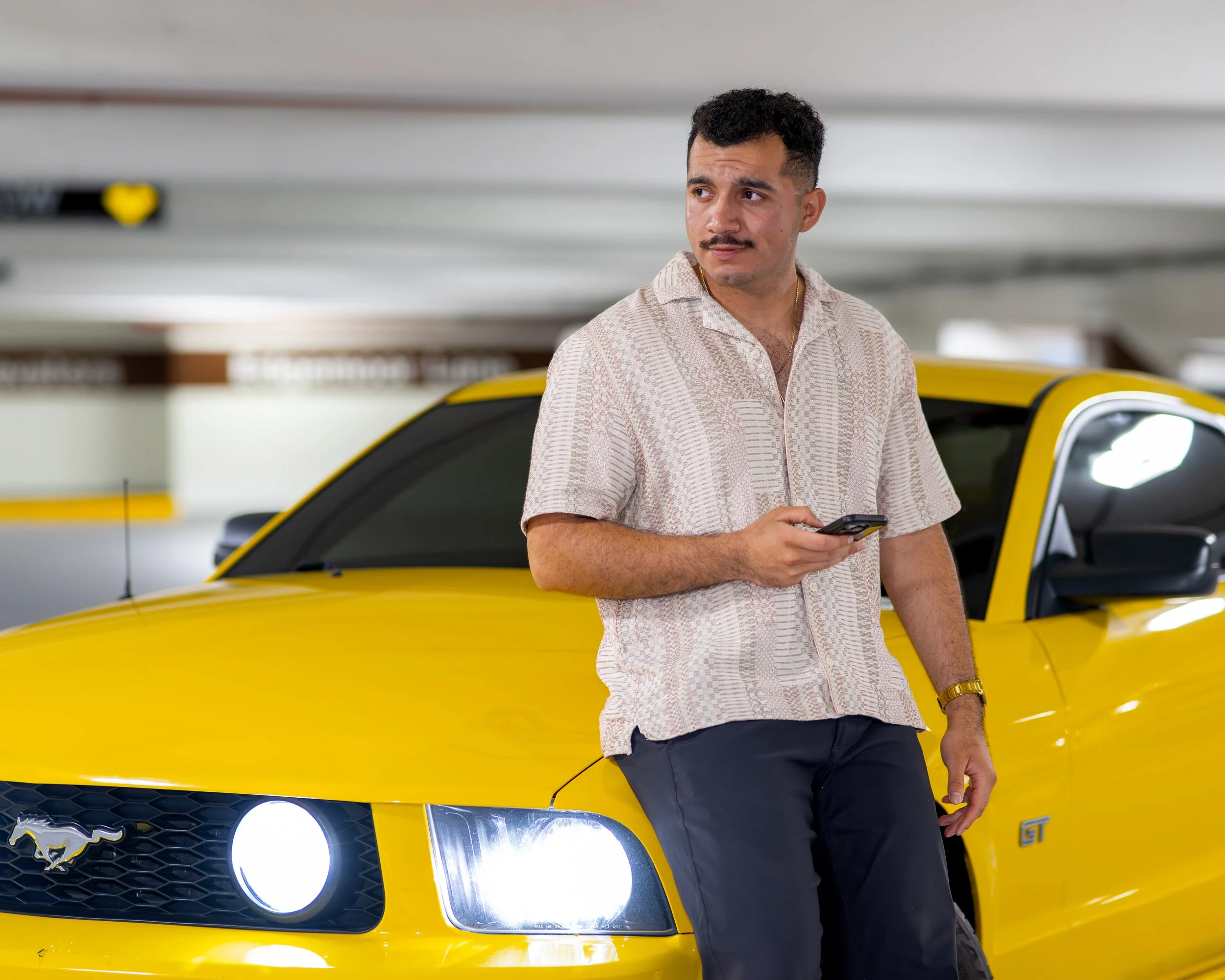 A man with dark hair and a mustache dressed in a light patterned shirt and dark pants, holding a smartphone, standing next to a yellow Ford Mustang GT in an indoor parking garage.