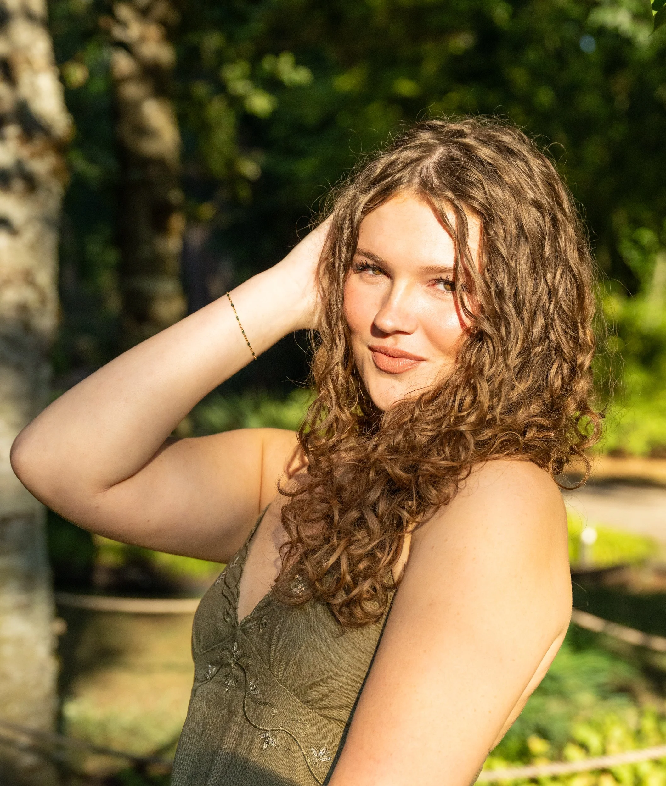 A senior portrait of a young woman with curly hair, wearing a sleeveless top with embroidery, poses outdoors against a backdrop of greenery and a trunk of a tree. She has a relaxed expression, with one hand touching her hair.