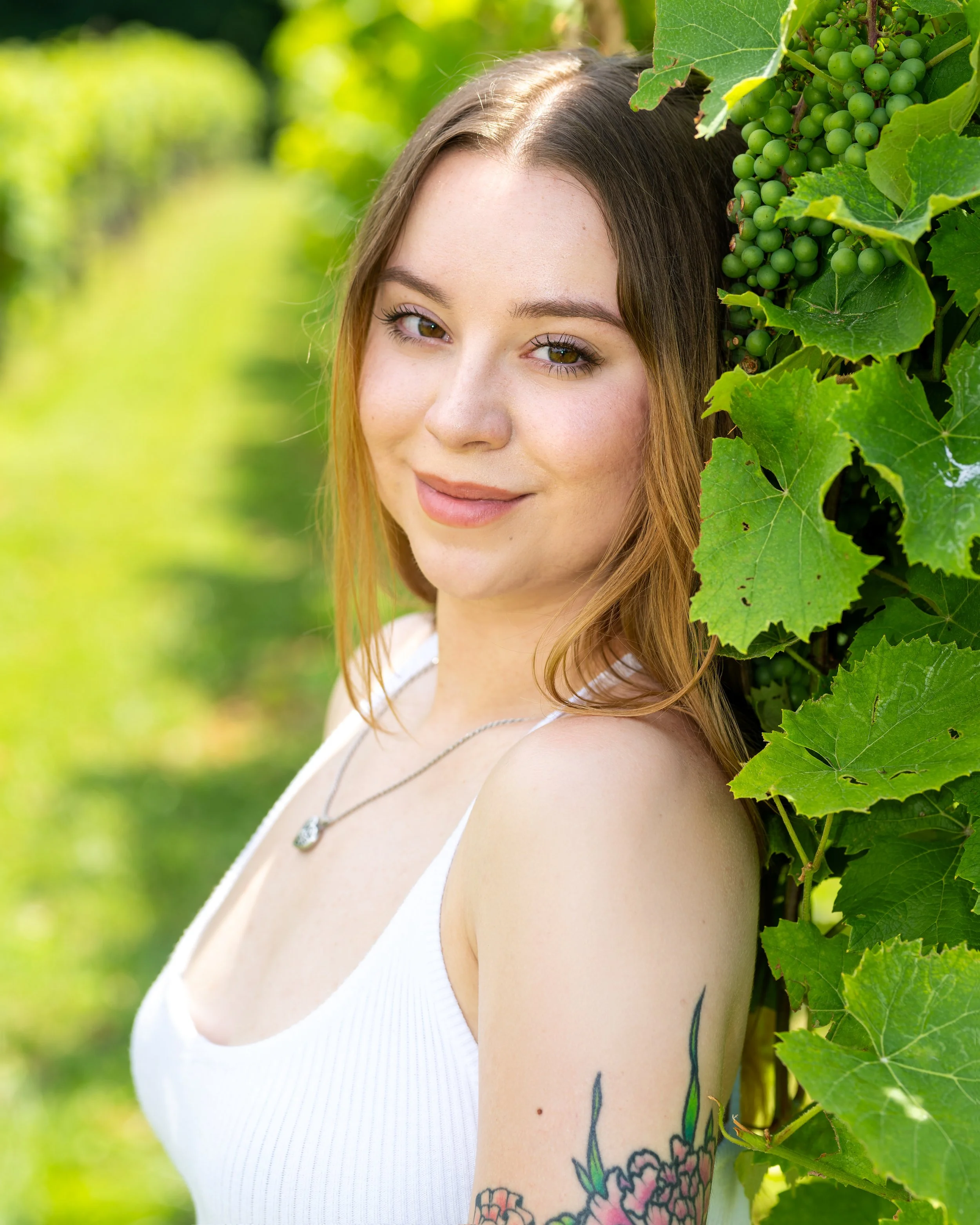 Close-up of a young woman with light brown hair, tattooed arm, wearing a white sleeveless top, and a silver necklace, standing outdoors among green grapevine leaves and bunches of unripe green grapes.