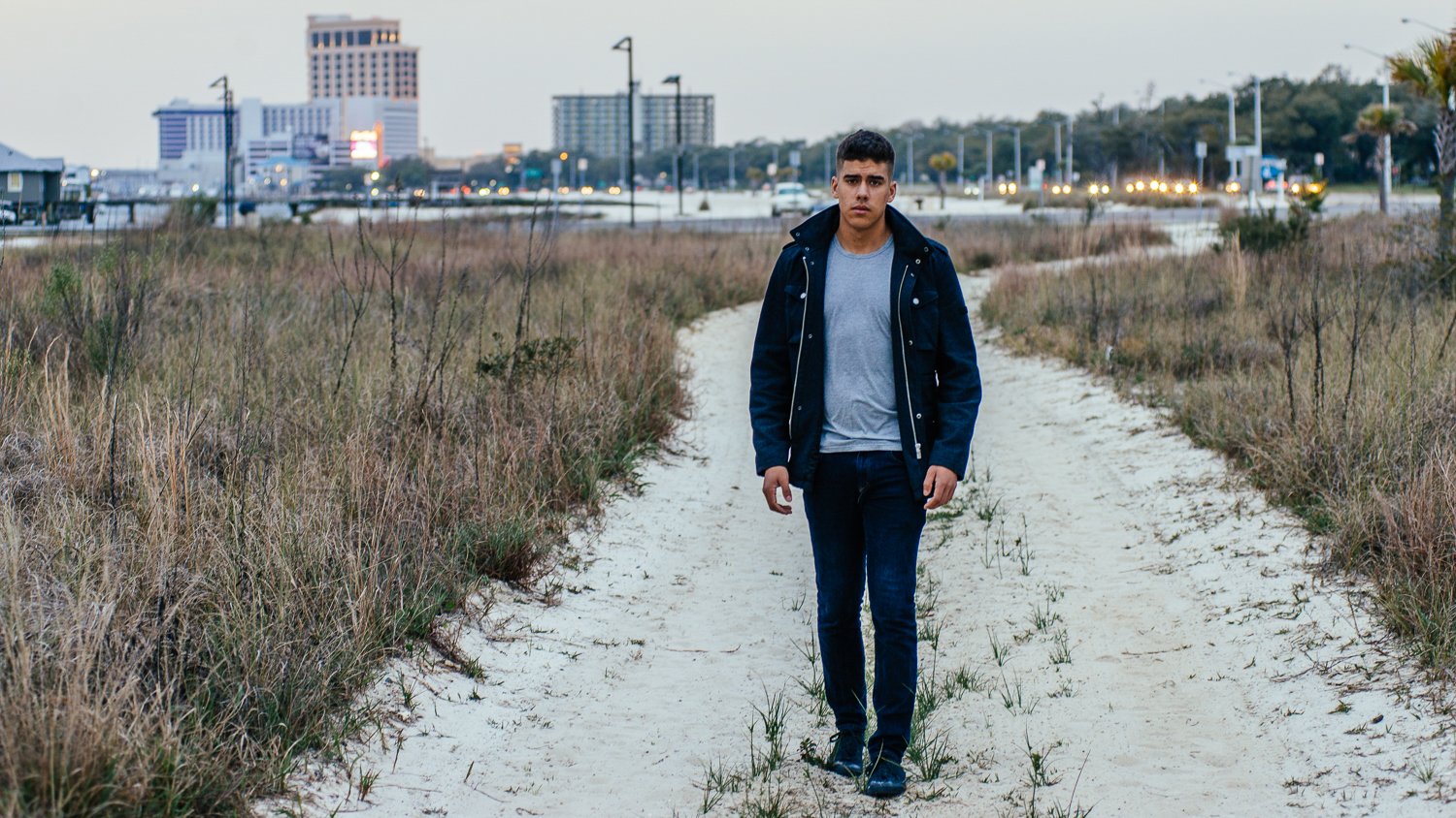 A young man walking on a sandy trail through dry grass in an urban area during early evening, with city buildings and lights in the background.