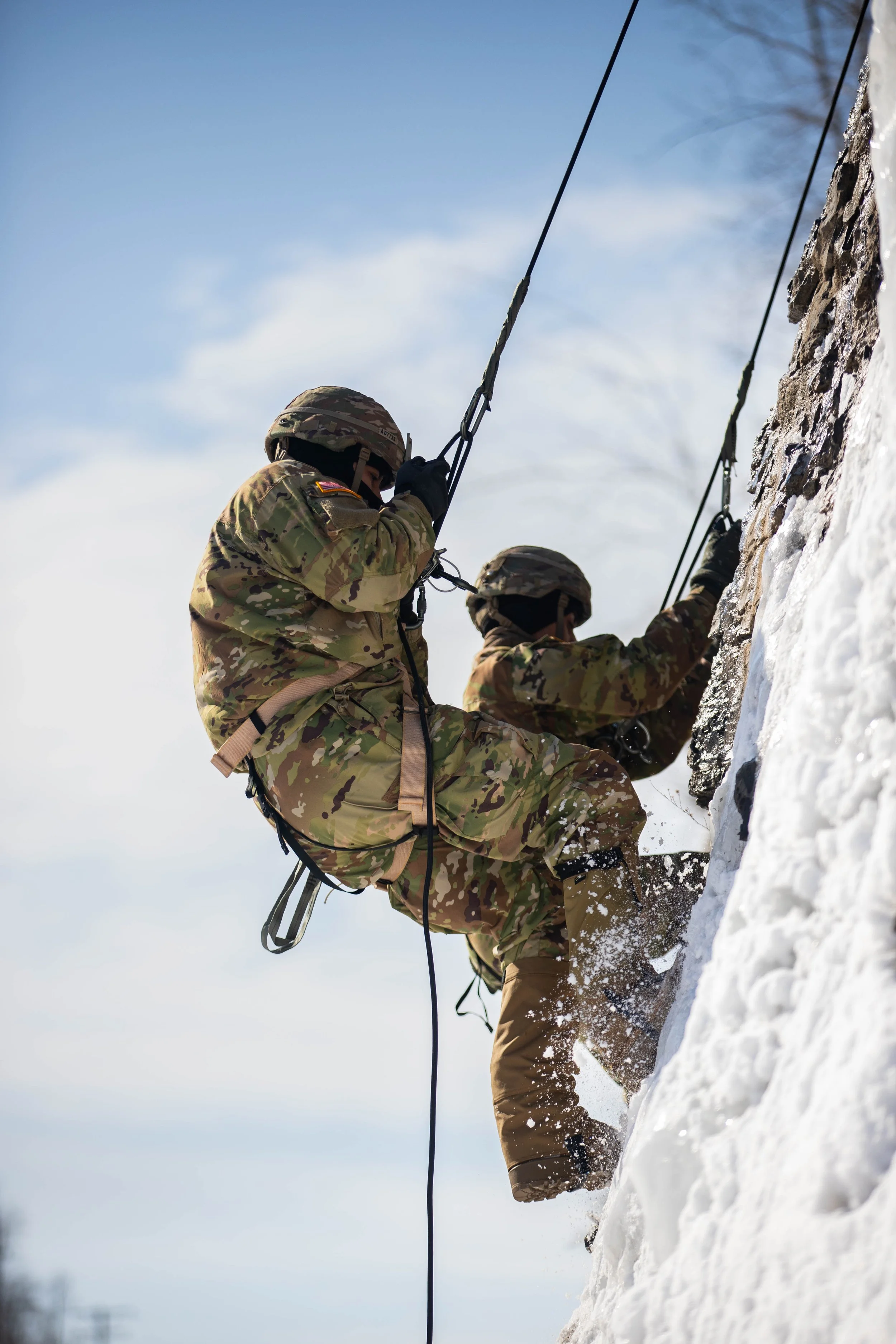 Two soldiers in camouflage uniforms and helmets climbing icy rock face with climbing gear, under clear sky.