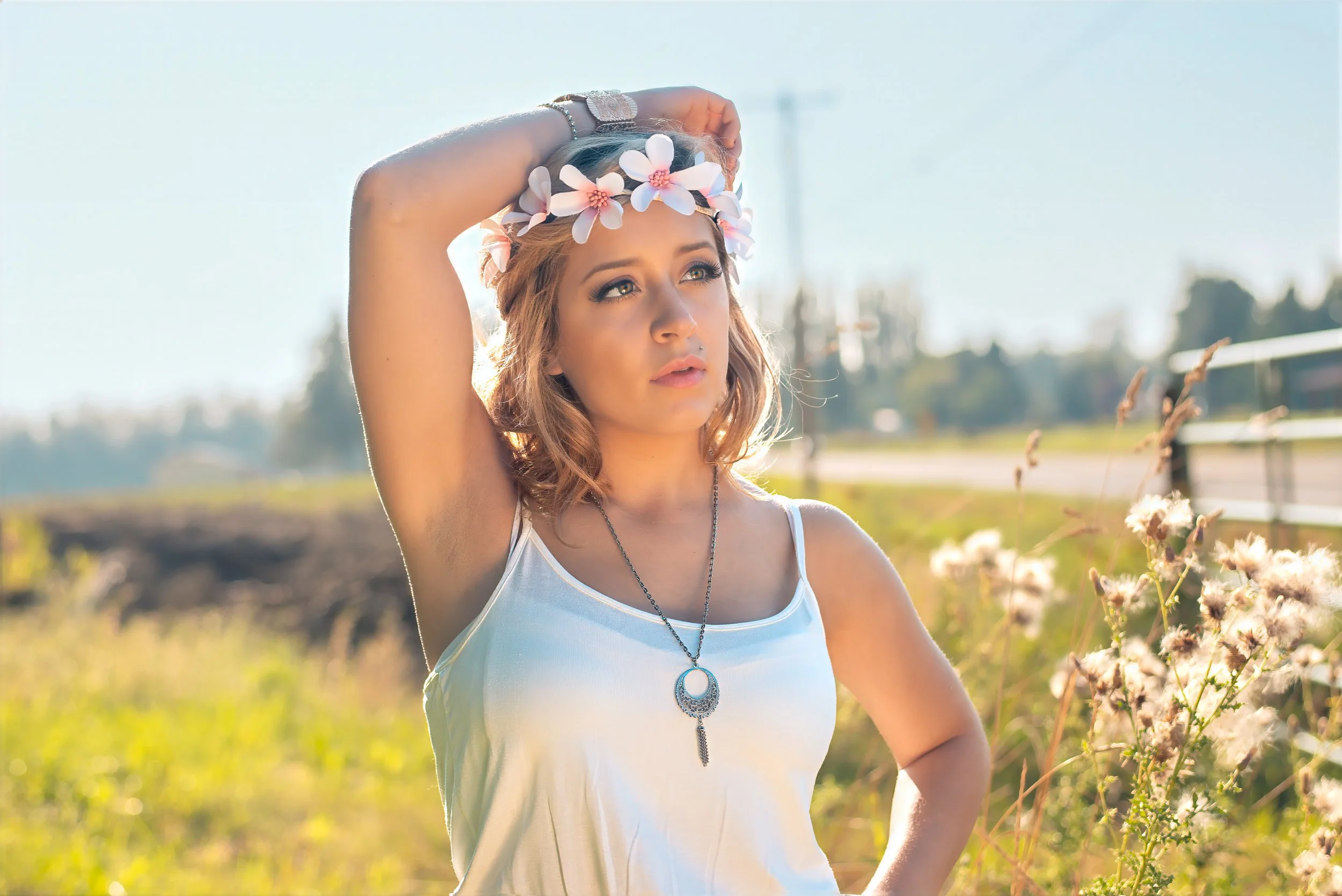 Young woman with long blonde hair wearing a white tank top, floral headband, and silver jewelry, standing outdoors in a field of flowers during daytime, gazing thoughtfully into the distance.