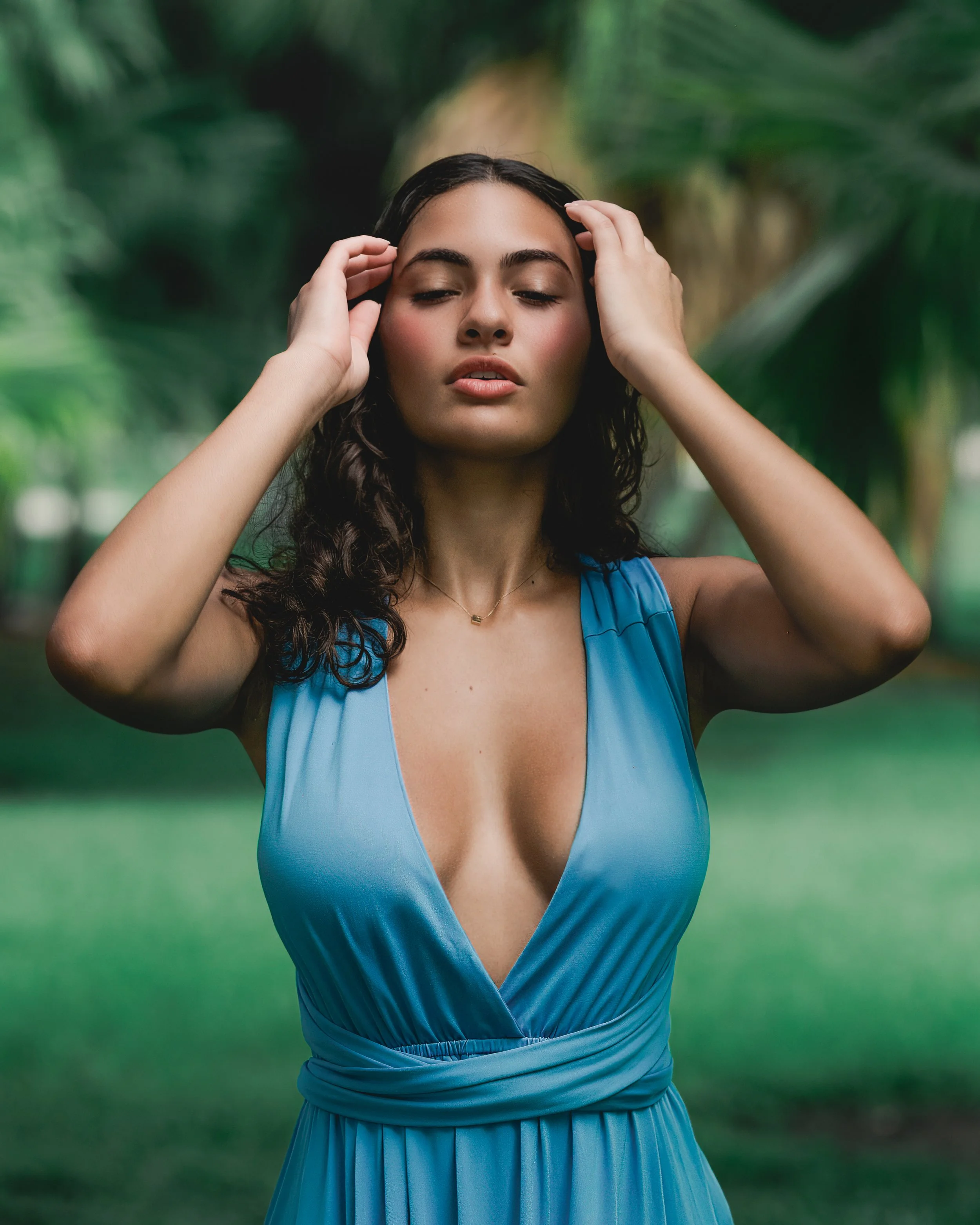 A woman with dark, wavy hair and light skin wearing a blue dress with a deep V neckline and a pleated skirt, standing outdoors with a lush green blurred background, her eyes closed and hands touching her head.