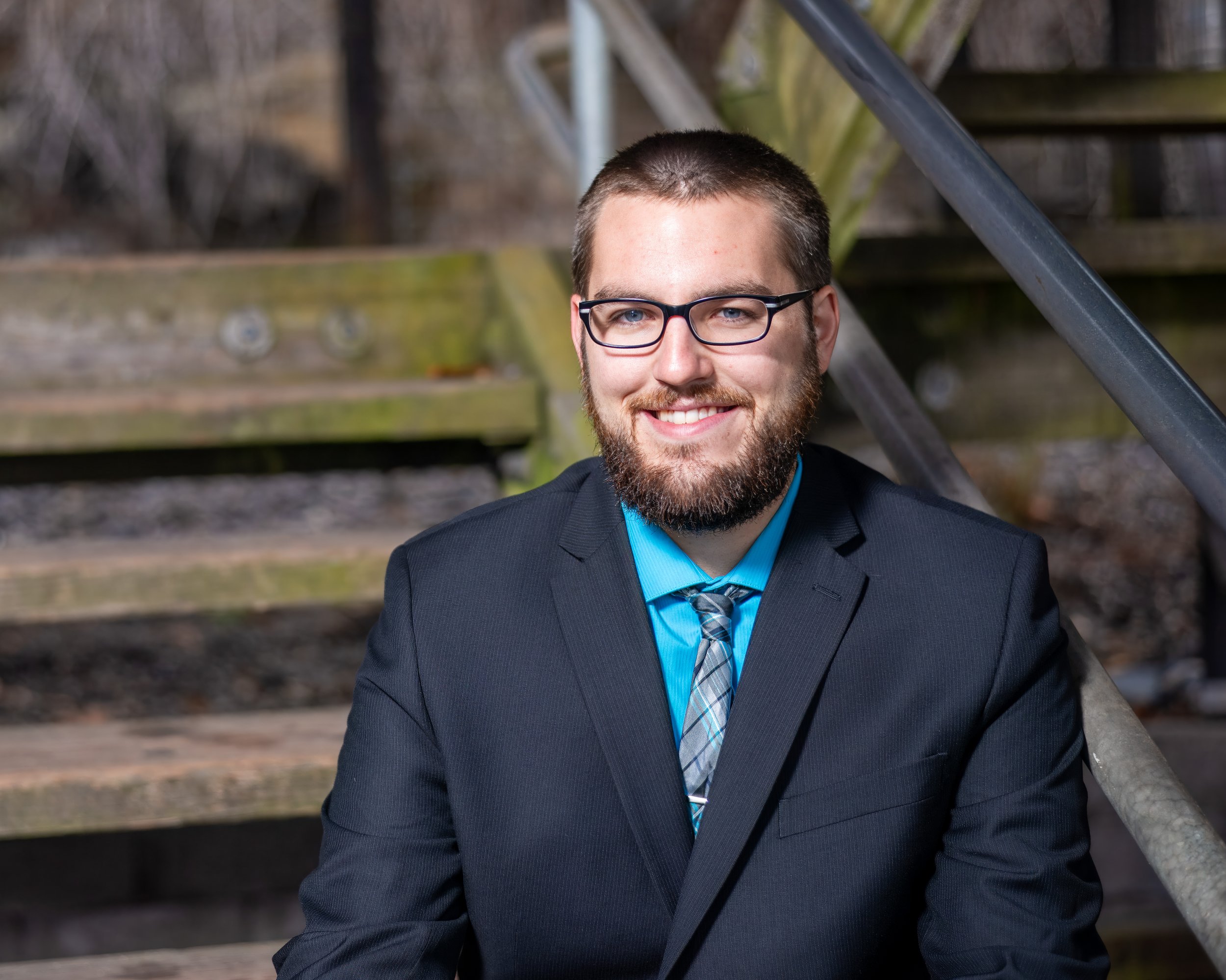 A man in a suit with a blue shirt and tie, wearing glasses, smiling outdoors in front of a wooden staircase.