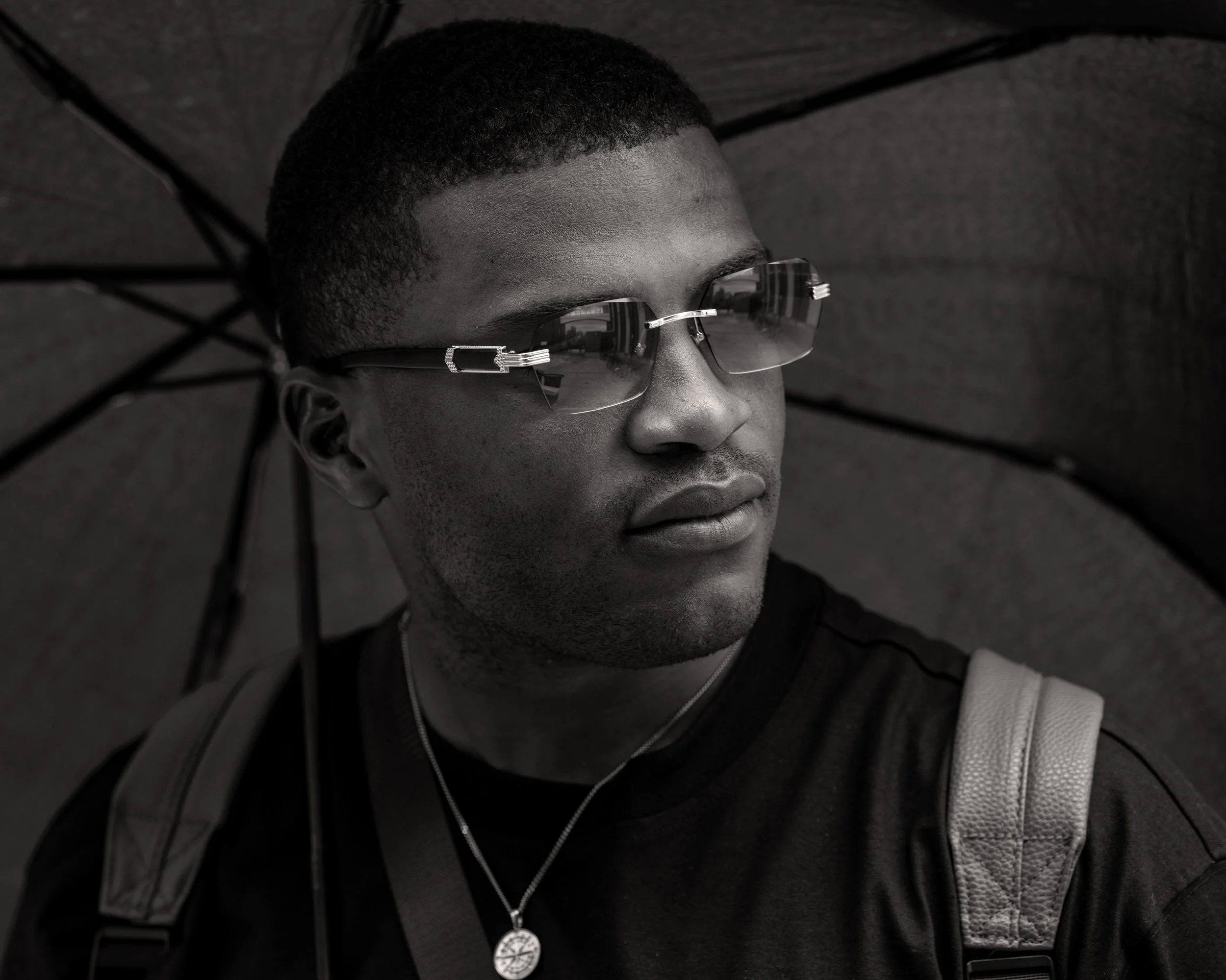 Close-up of a man with short curly hair wearing sunglasses, a necklace with a circular pendant, and a dark zip-up jacket, lying on a dark textured surface.
