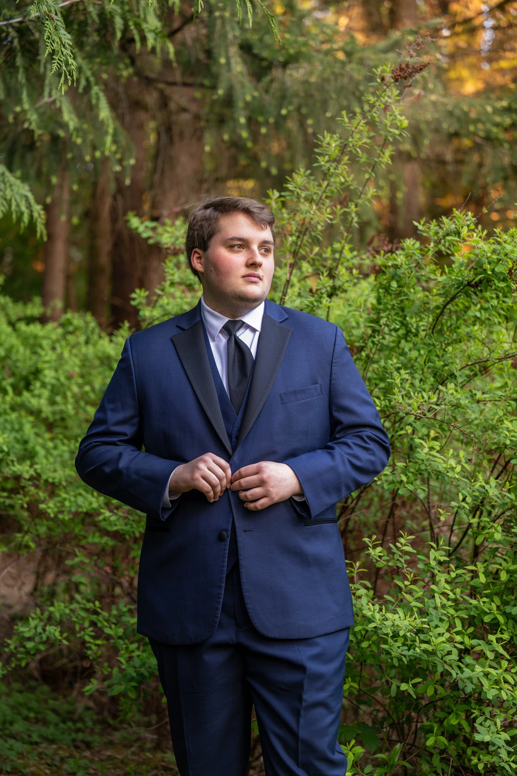 A young man dressed in a navy blue suit and tie stands outdoors among lush green bushes and trees, looking off into the distance with a contemplative expression.