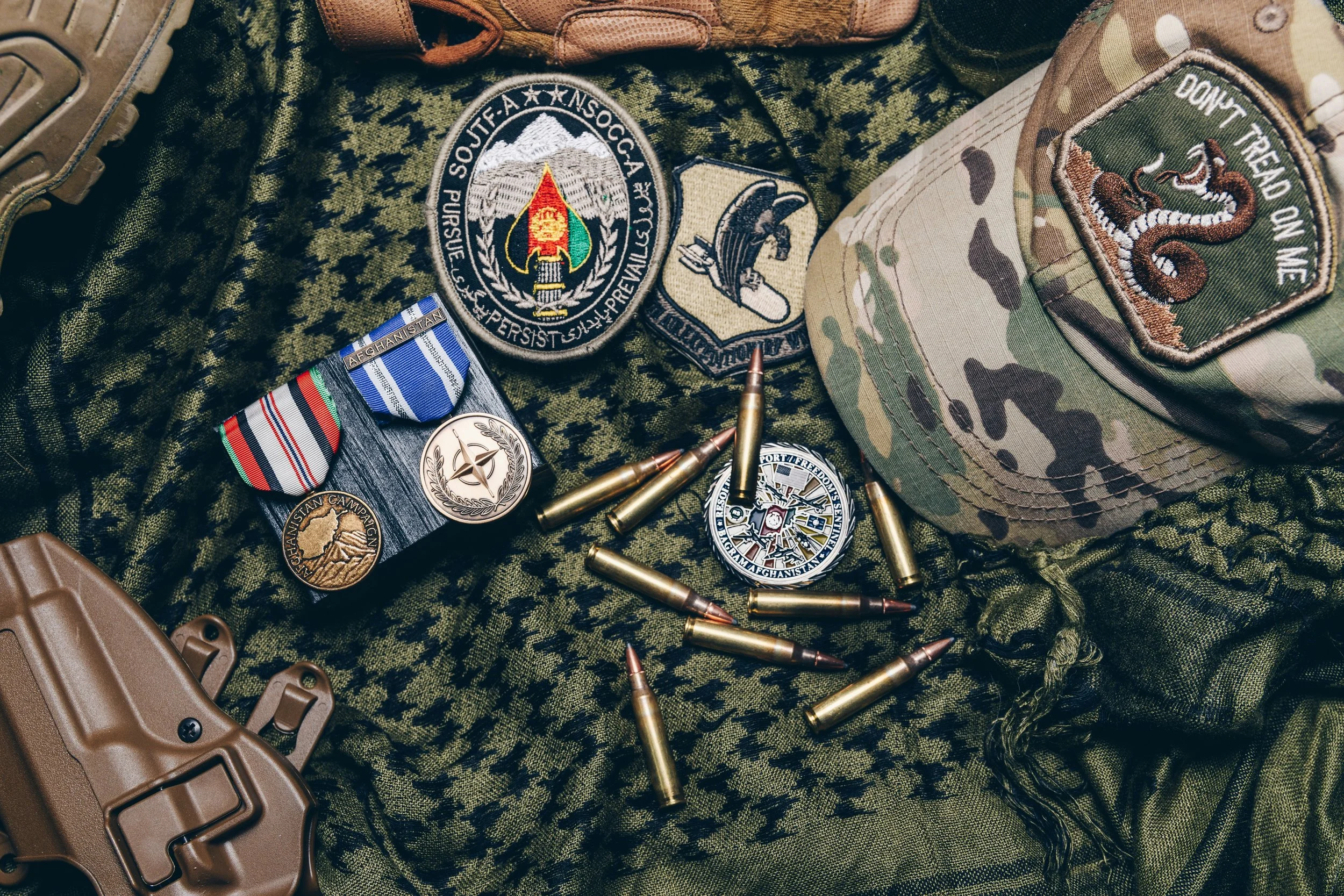 Military patches, medals, bullets, and a tan tactical helmet on camouflage fabric that reads 'DON'T TREAD ON ME' with a coiled snake patch.