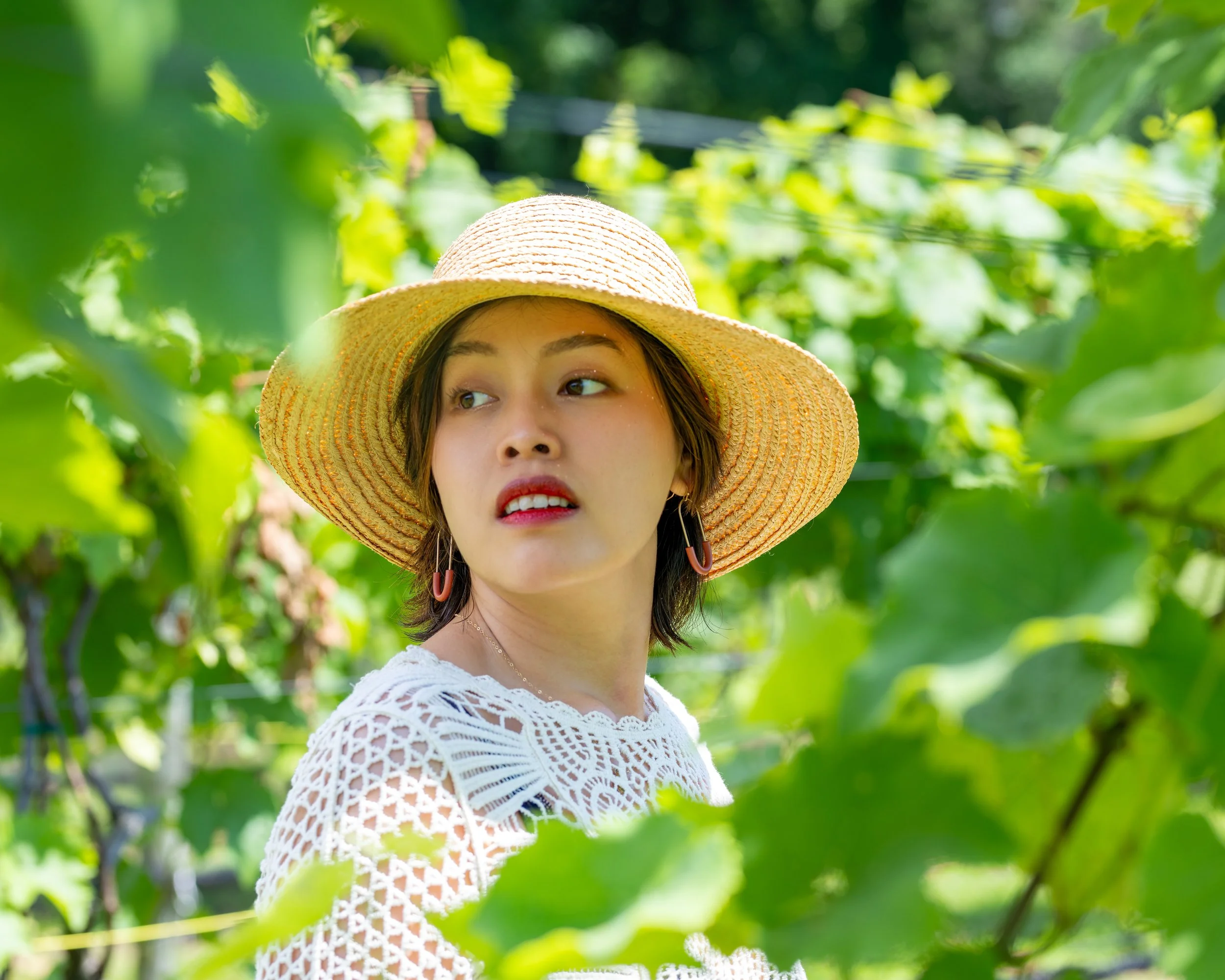 A woman wearing a wide-brimmed straw hat with a beige band, a white lace top, and earrings, standing among lush green foliage and vine plants, looking to the side.