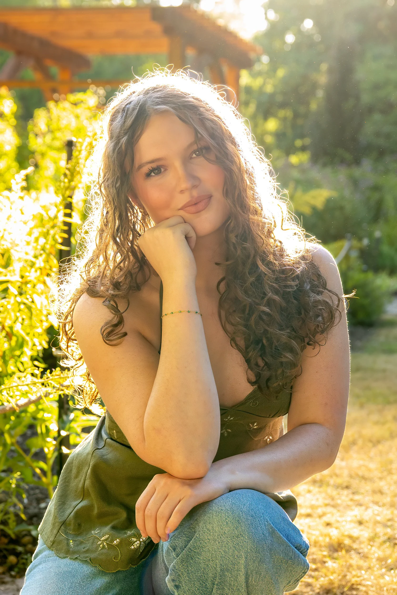 A young woman with long curly brown hair and light skin sitting outdoors in sunlight. She is resting her chin on her hand, smiling softly, with green trees and a wooden structure in the background.