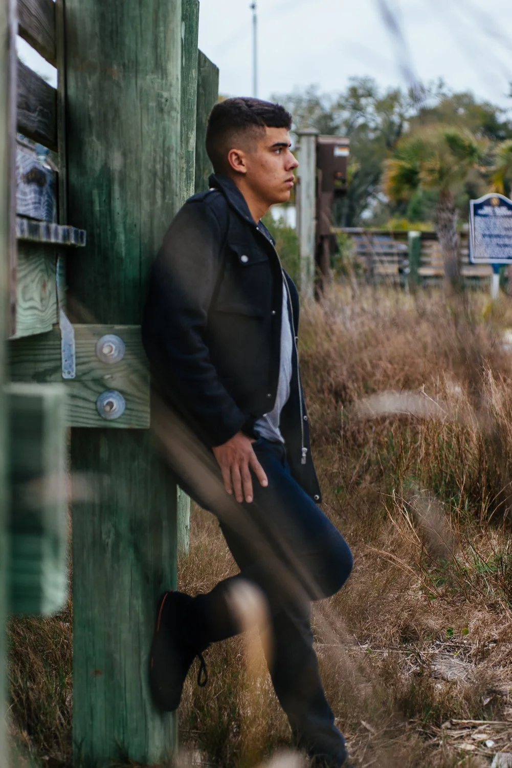 Young man with dark hair wearing a black jacket and jeans leaning against a weathered green wooden fence in a grassy outdoor area with trees and a blue informational sign in the background.