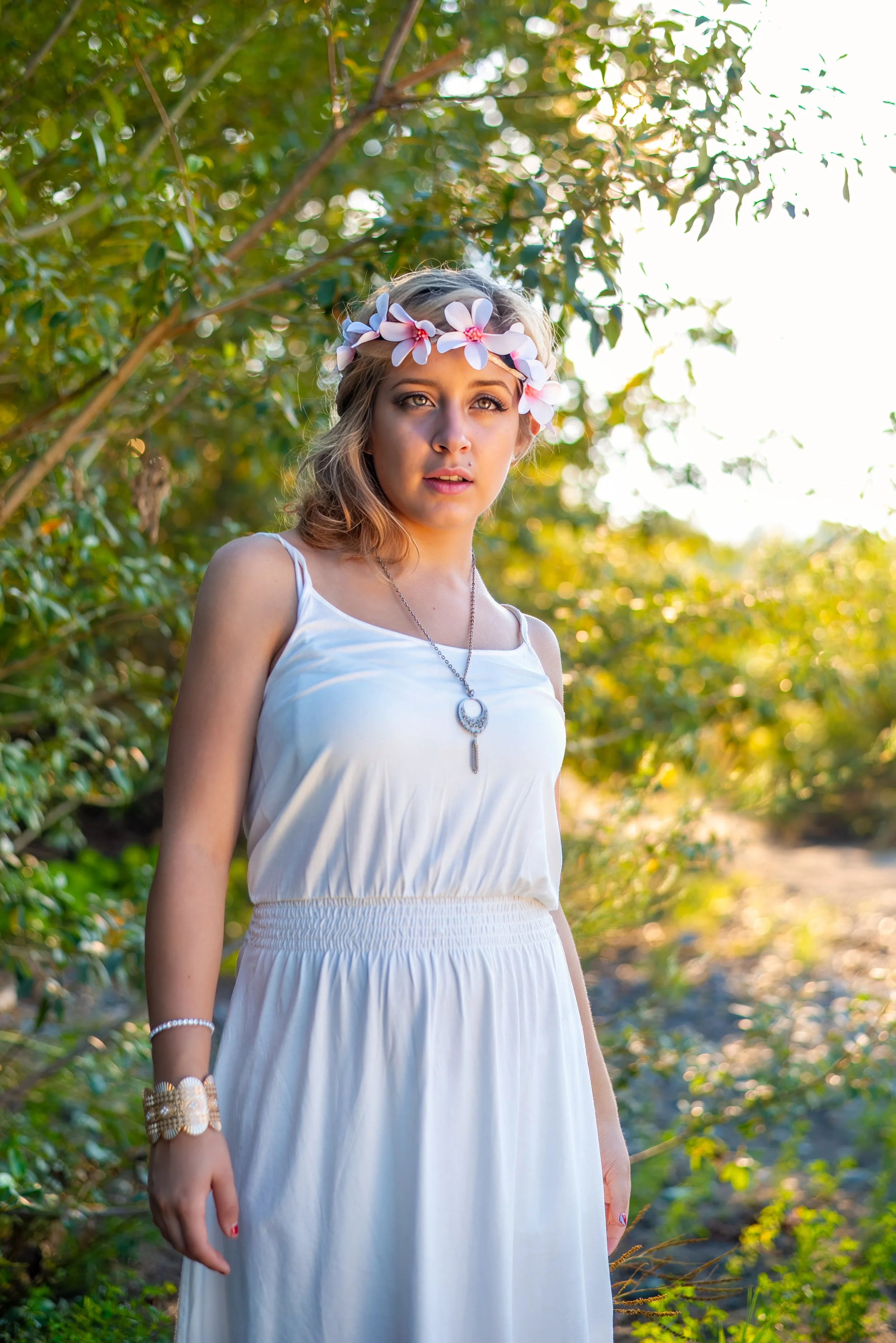 Young woman with blond hair, wearing a floral headband and white dress, standing outdoors in a lush green setting with sunlight filtering through the trees.