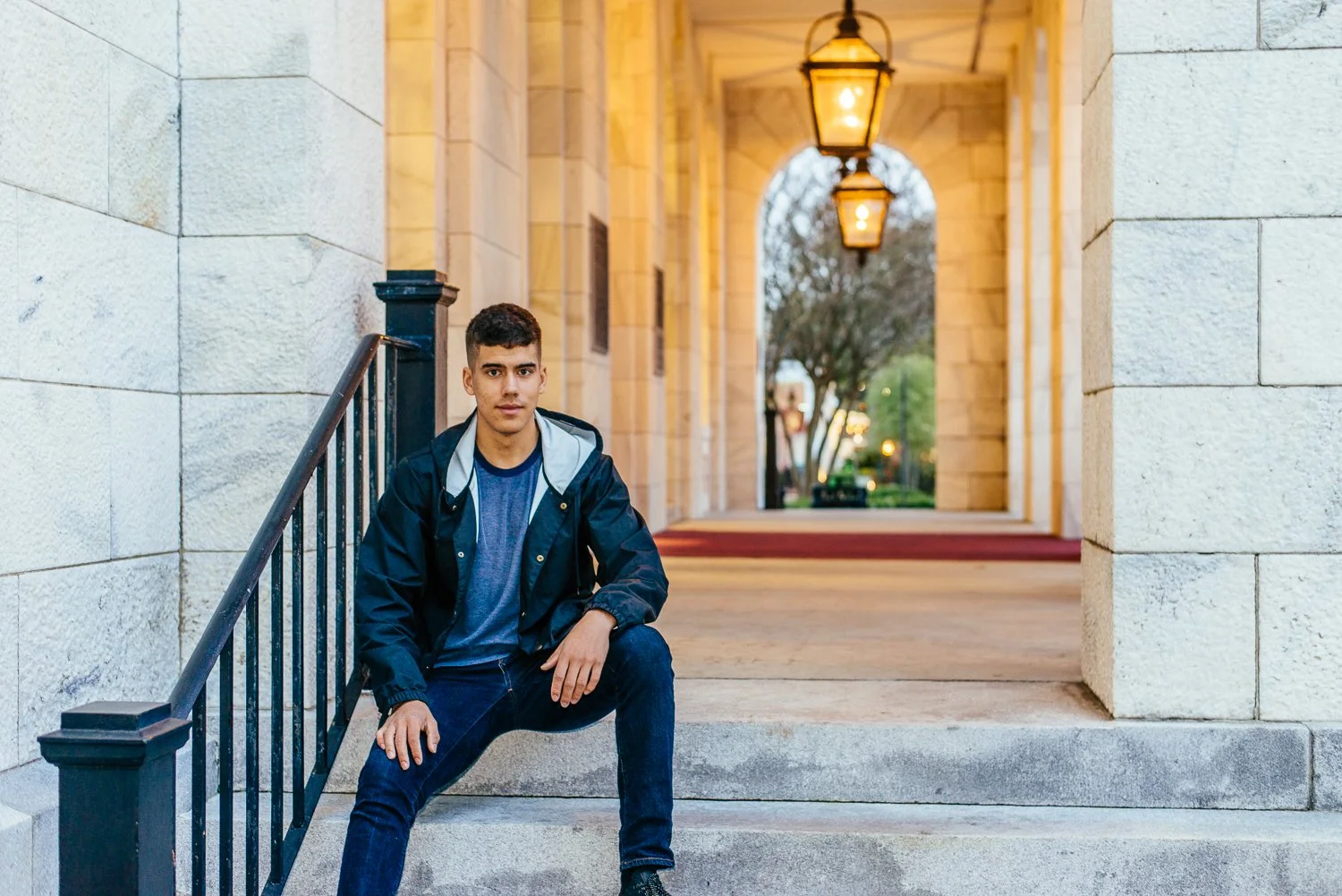 A young man in a blue jacket and jeans sitting on stairs under a stone archway, with a row of hanging lamps and trees in the background during dusk.