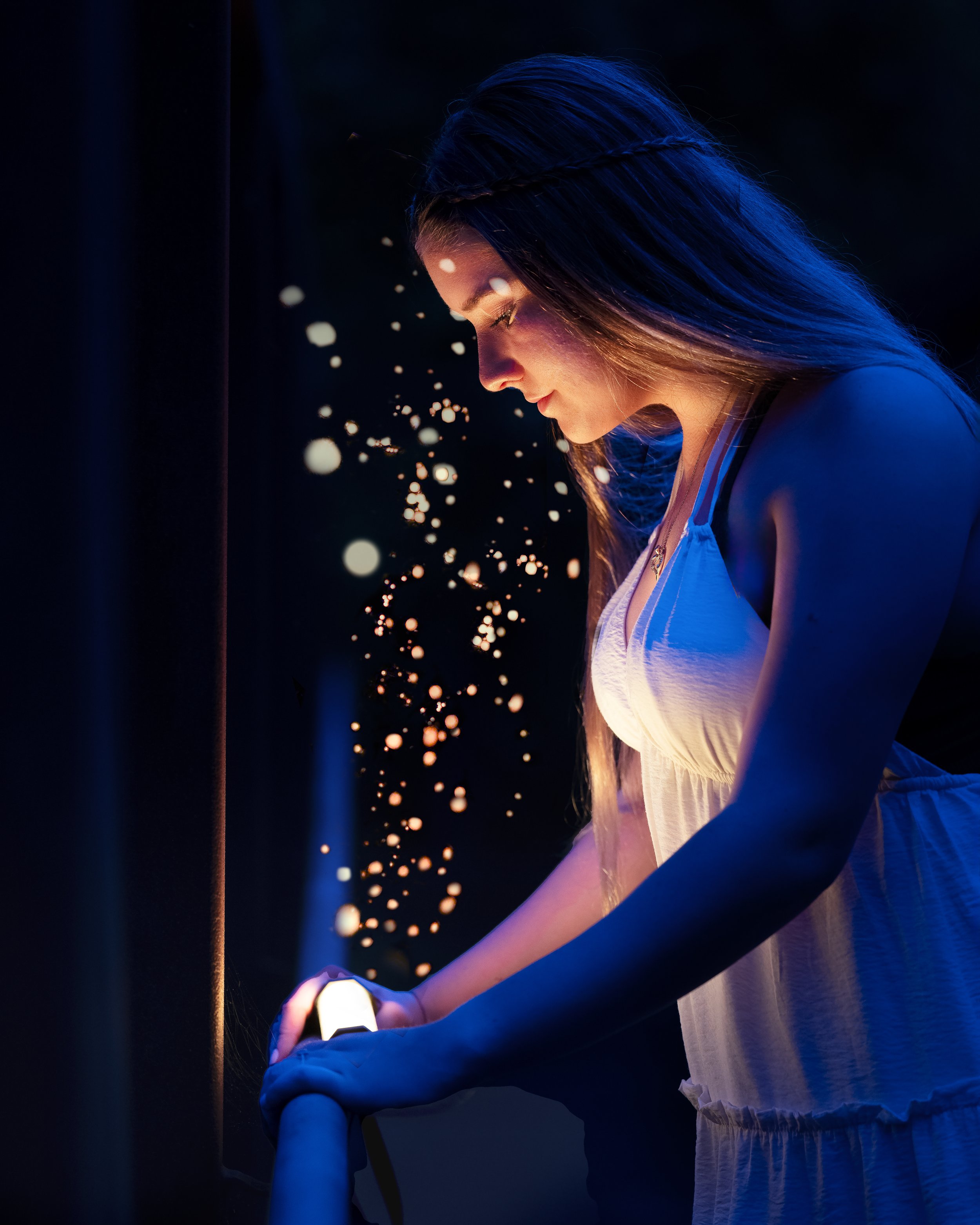 A senior portrait of a young woman with long hair in a braid, illuminated by blue and warm lighting, is looking at and interacting with an electronic device while sitting in a dark environment with sparks or glowing particles around her.