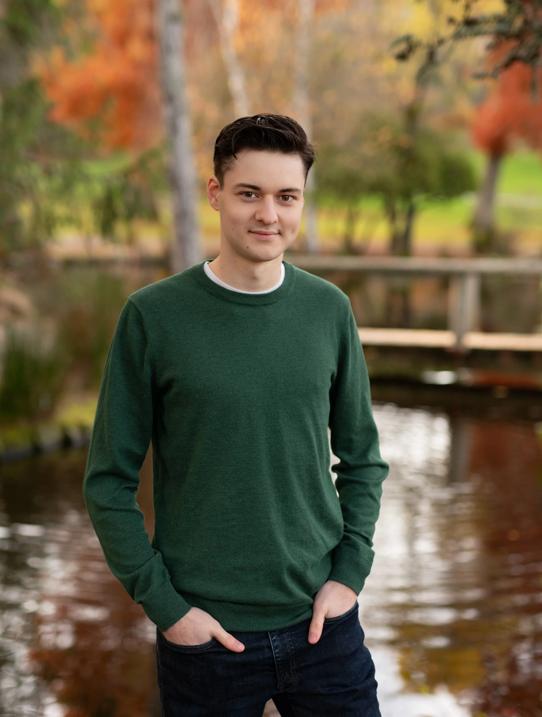 A young man with dark hair wearing a green sweater and blue jeans standing outdoors by a lake during fall, with trees showing autumn colors in the background.