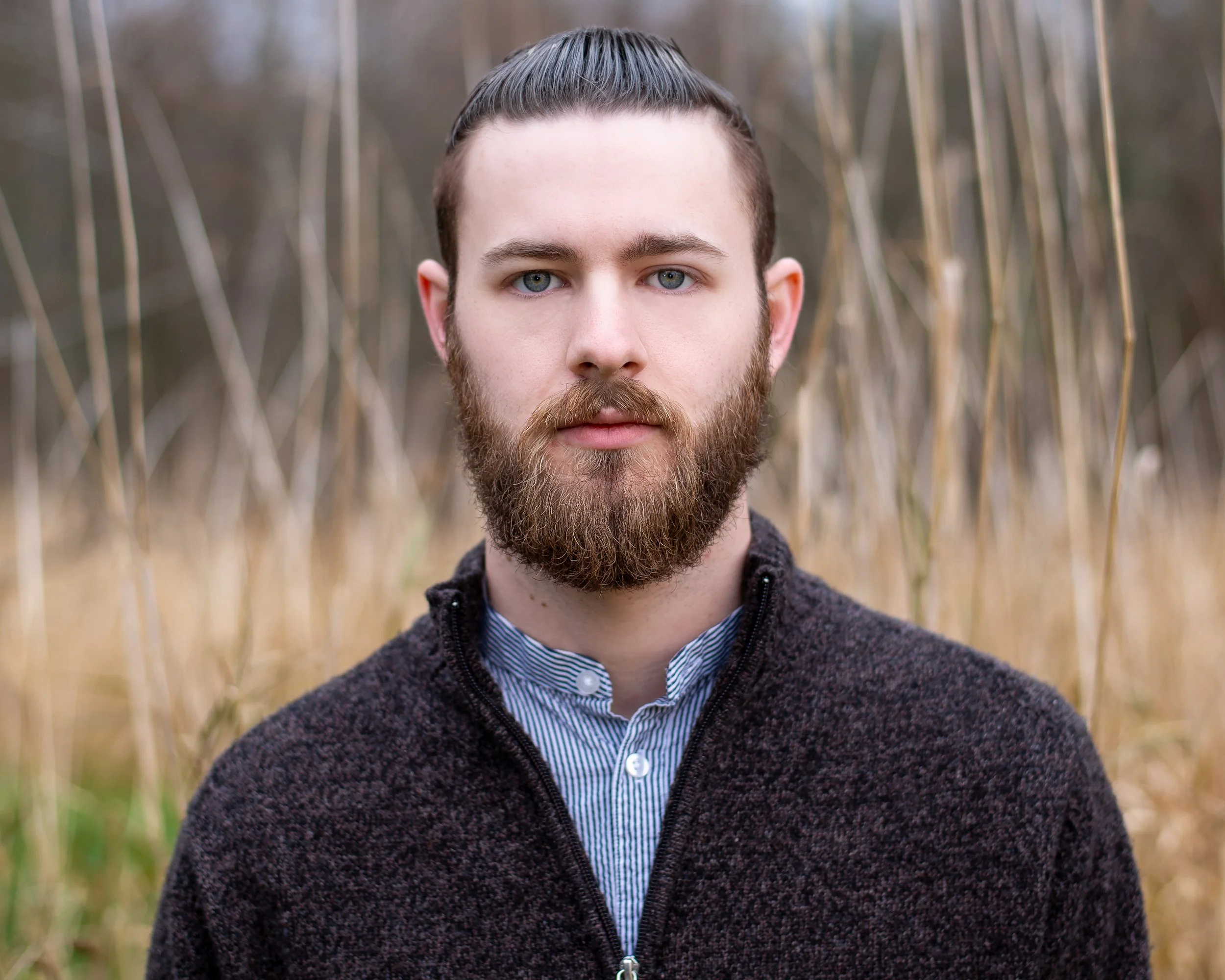 Close-up of a young man with a beard, blue eyes, and dark brown hair styled back, standing outdoors in a field of tall, dry grass, wearing a dark, zip-up sweater over a blue and white striped shirt.