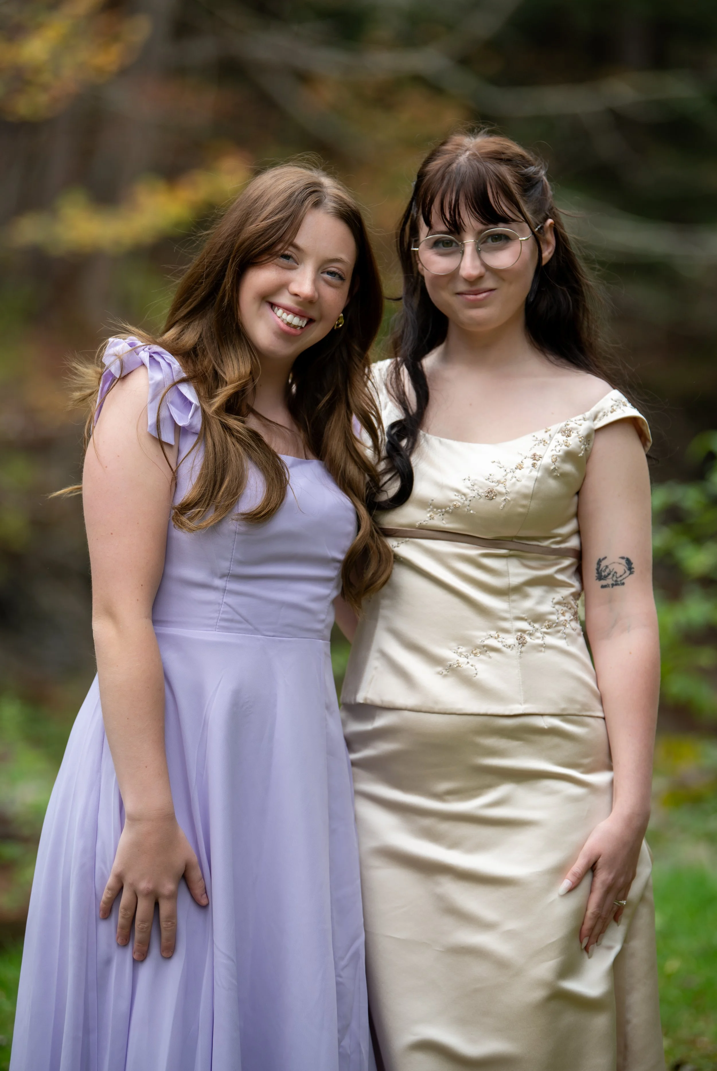 Two women in elegant dresses standing close together outdoors with a blurred natural background.