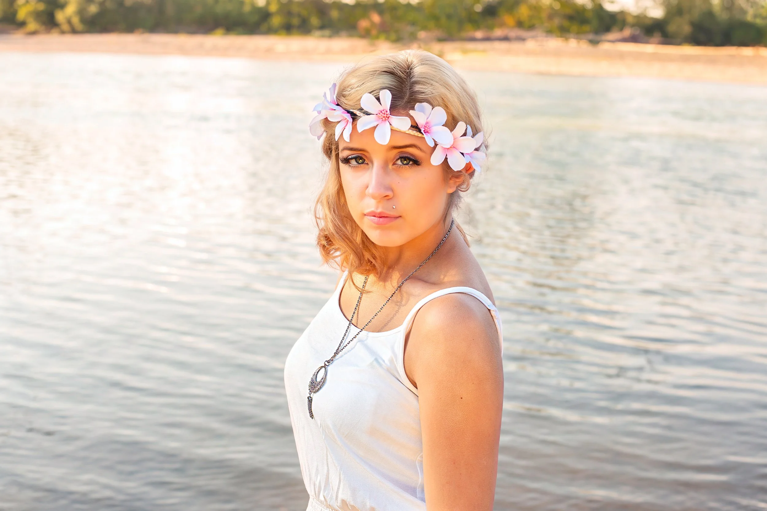 Young woman with blonde hair wearing a white top and floral crown, standing near water with trees in the background.