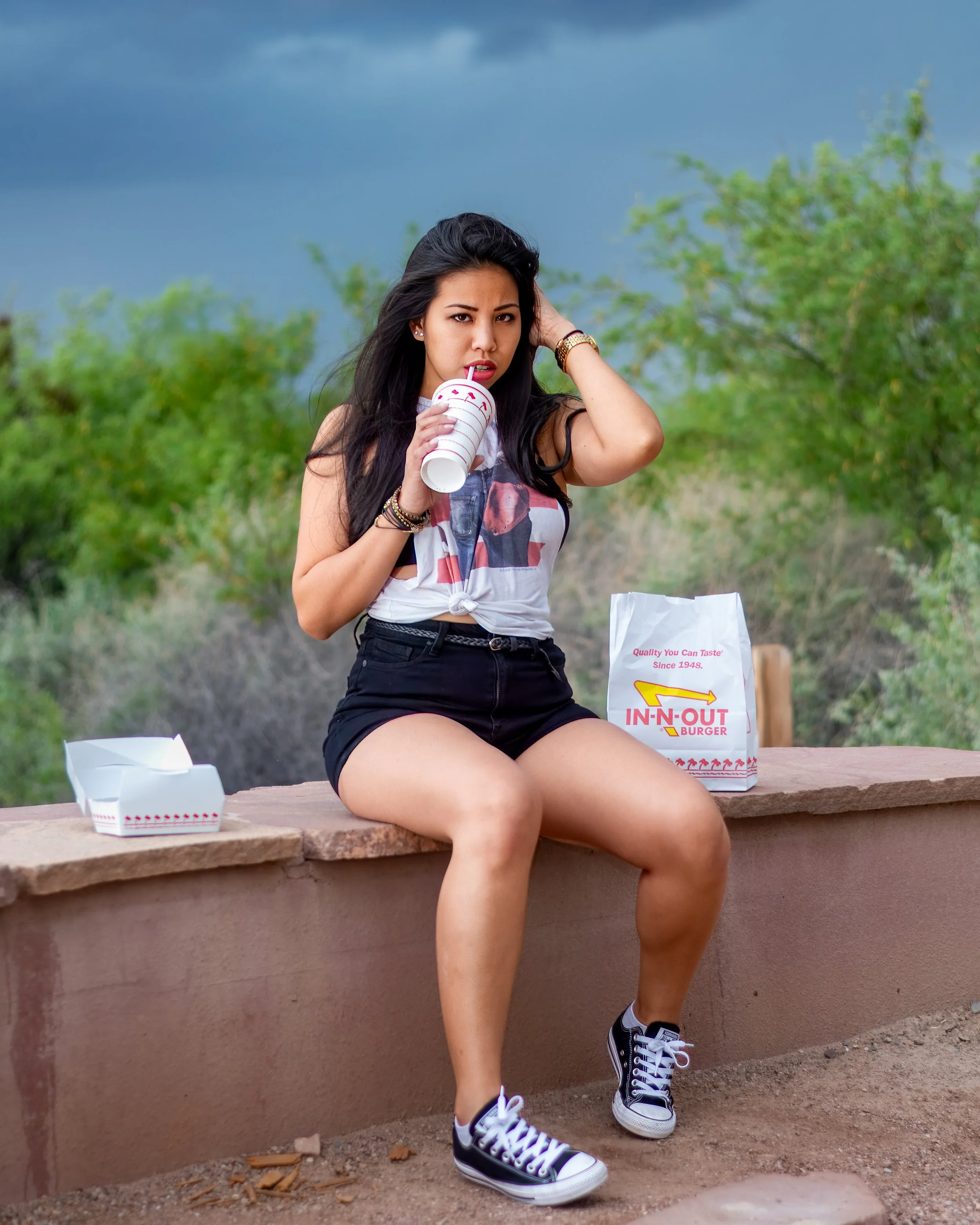Young woman sitting on a low wall outside, drinking from a paper cup with a straw, surrounded by fast food containers, with a cloudy sky and greenery in the background.