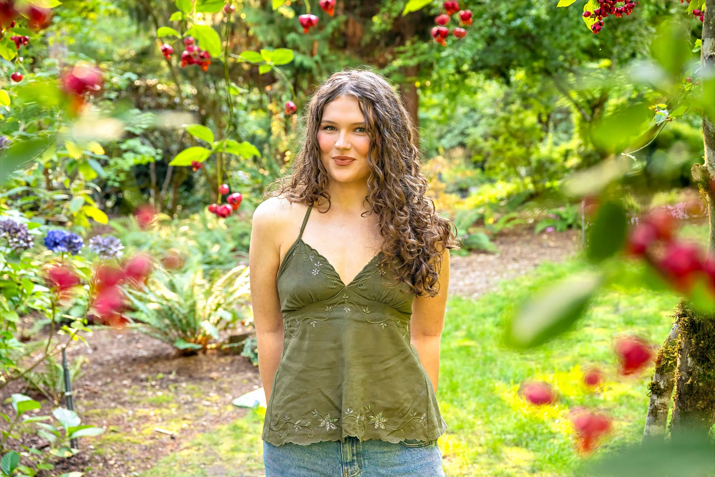 A senior portrait of a woman with long, curly brown hair standing outdoors in a garden with green trees and plants around her, wearing a green embroidered sleeveless top and blue jeans.