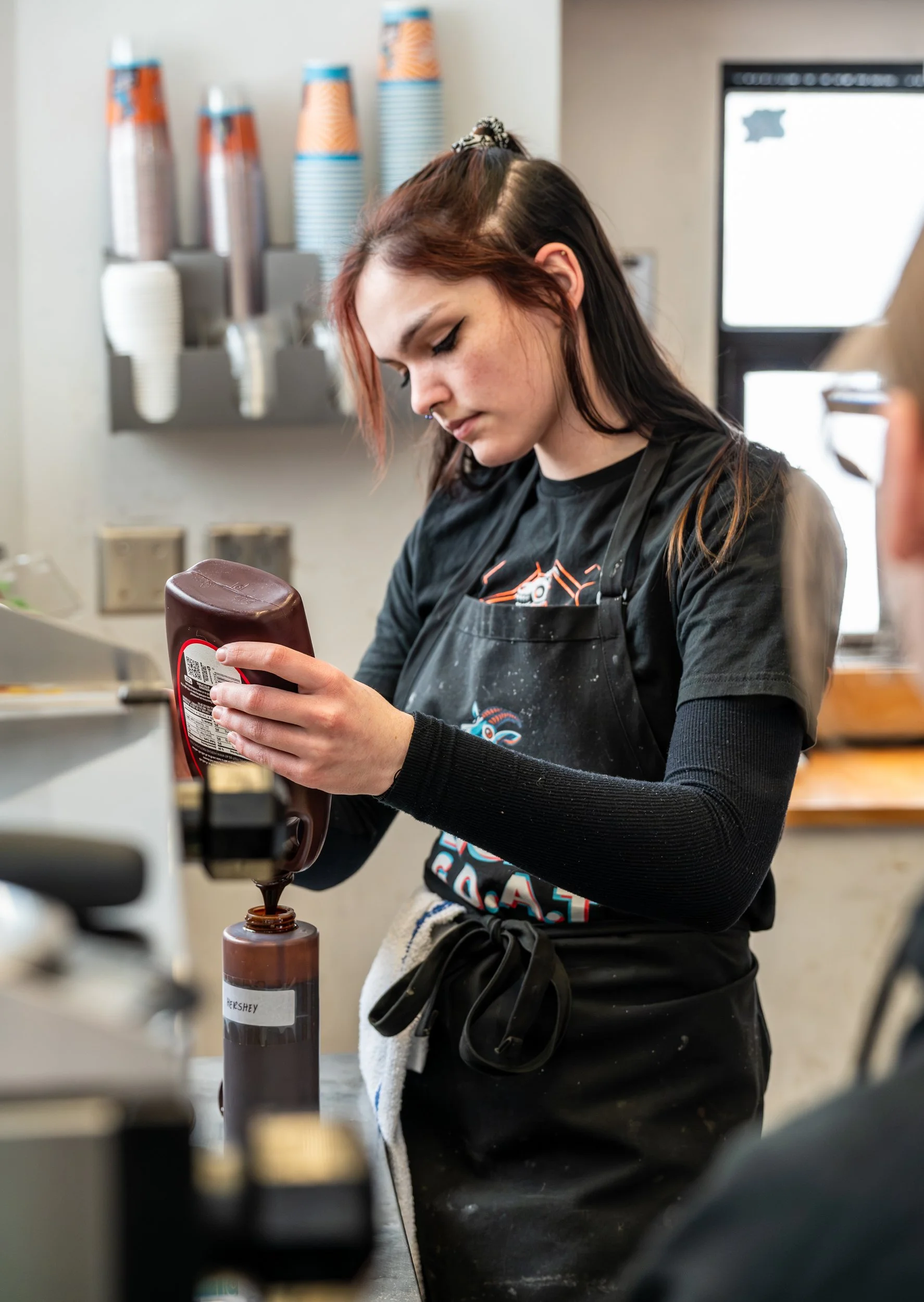 Young woman with dark and red hair, wearing a black apron, pouring chocolate syrup from a bottle onto a dessert, with a background of soda cups and a drink machine.