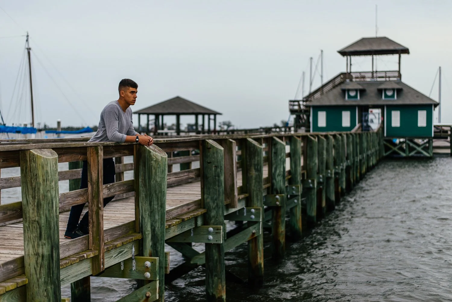 A young man leaning on a wooden dock by the water, looking pensively into the distance with boats and a green building in the background.