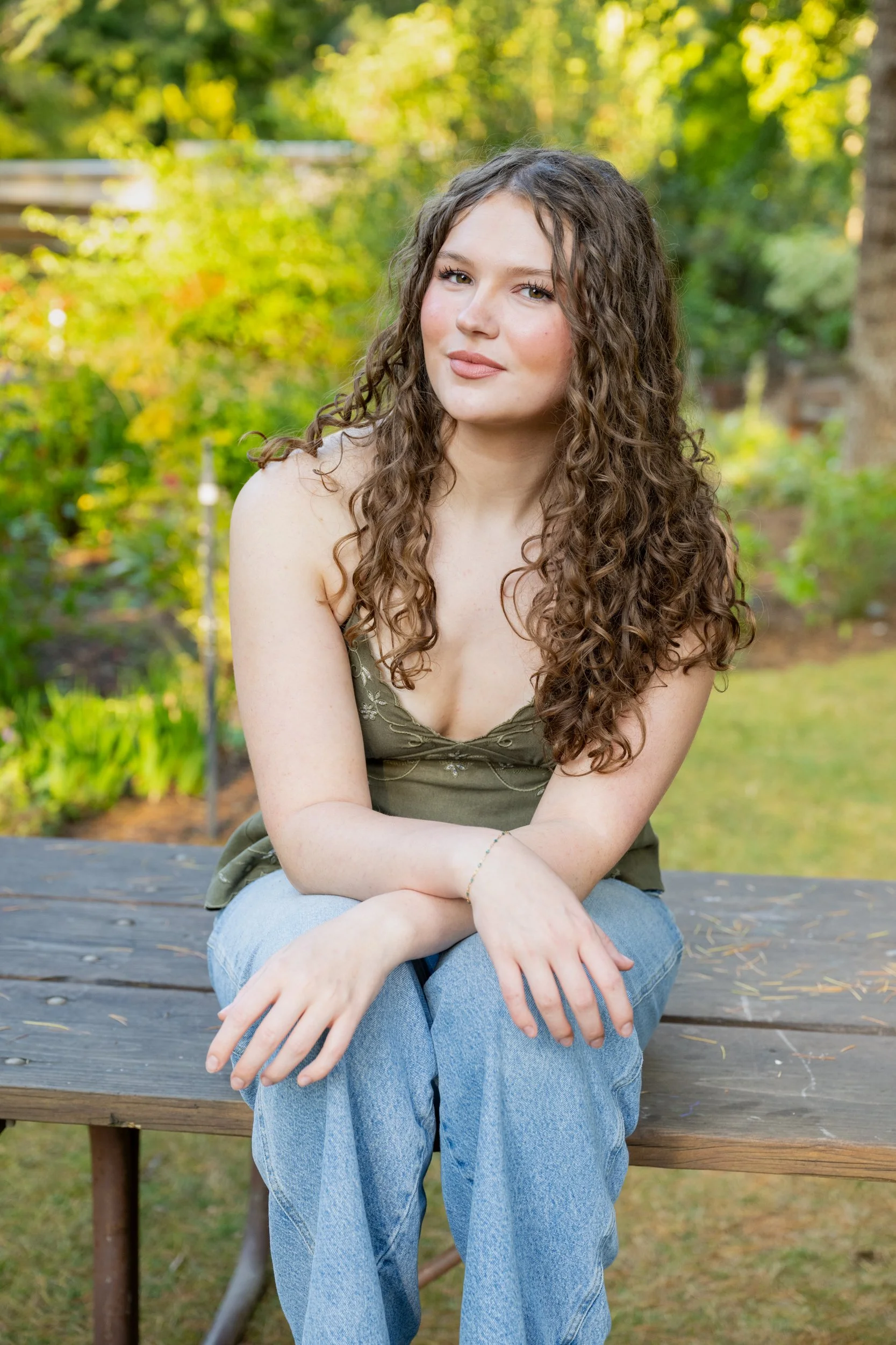 A senior portrait of a young woman with long, curly hair sitting outdoors on a wooden bench, wearing a green top and jeans, with a background of green trees and sunlight.