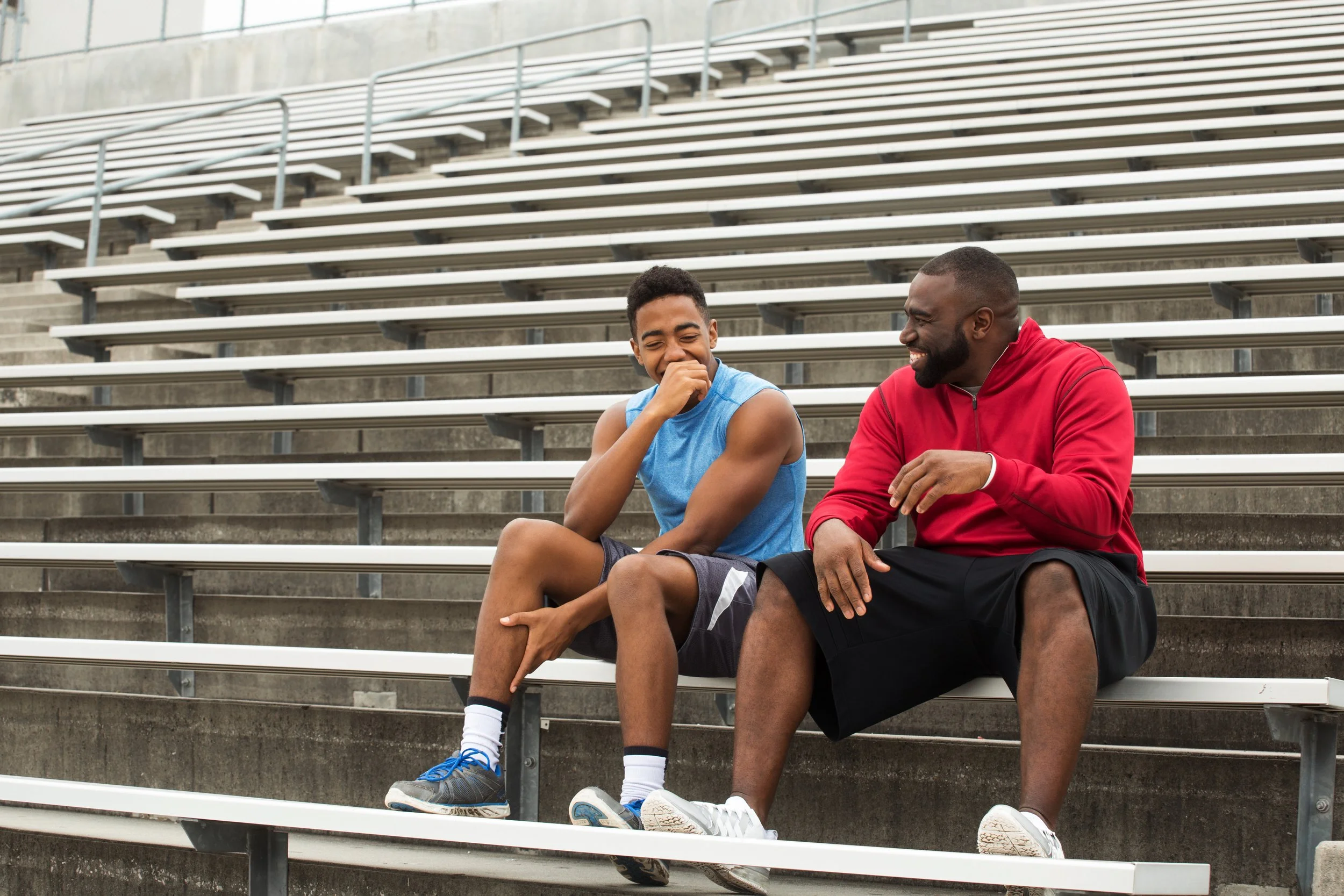 A young man and an older man sitting on stadium bleachers, laughing and talking; the young man in athletic gear and the older man in a red hoodie and black shorts.