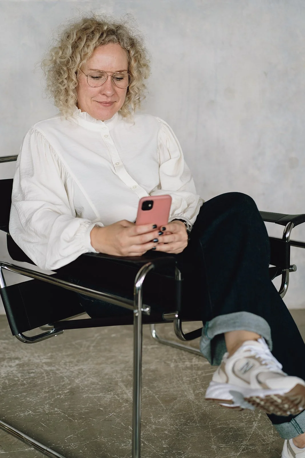 A woman with blonde curly hair, wearing glasses and a white blouse, sitting in a modern black chair, looking at her pink smartphone, with her legs crossed, in a minimalist indoor setting.