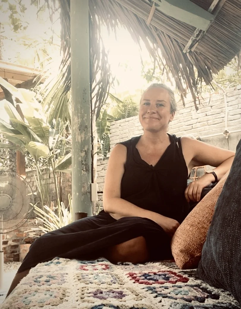 A woman in a black sleeveless top sitting on a colorful, patterned rug under a thatched roof, with greenery and a brick wall in the background, smiling.