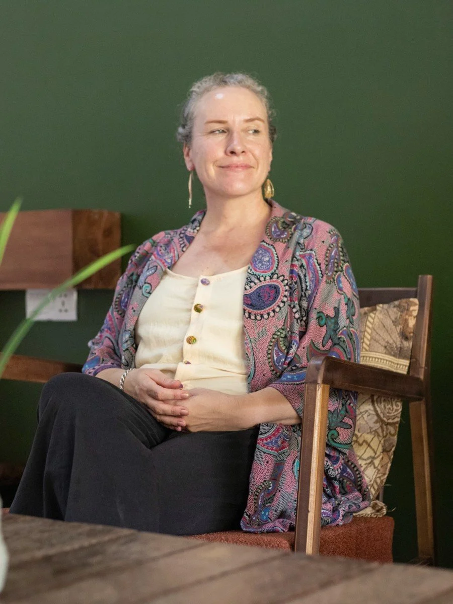 A woman with curly blonde hair sitting on a sofa, holding a white mug, looking thoughtfully to the side, wearing a black shirt and a watch.