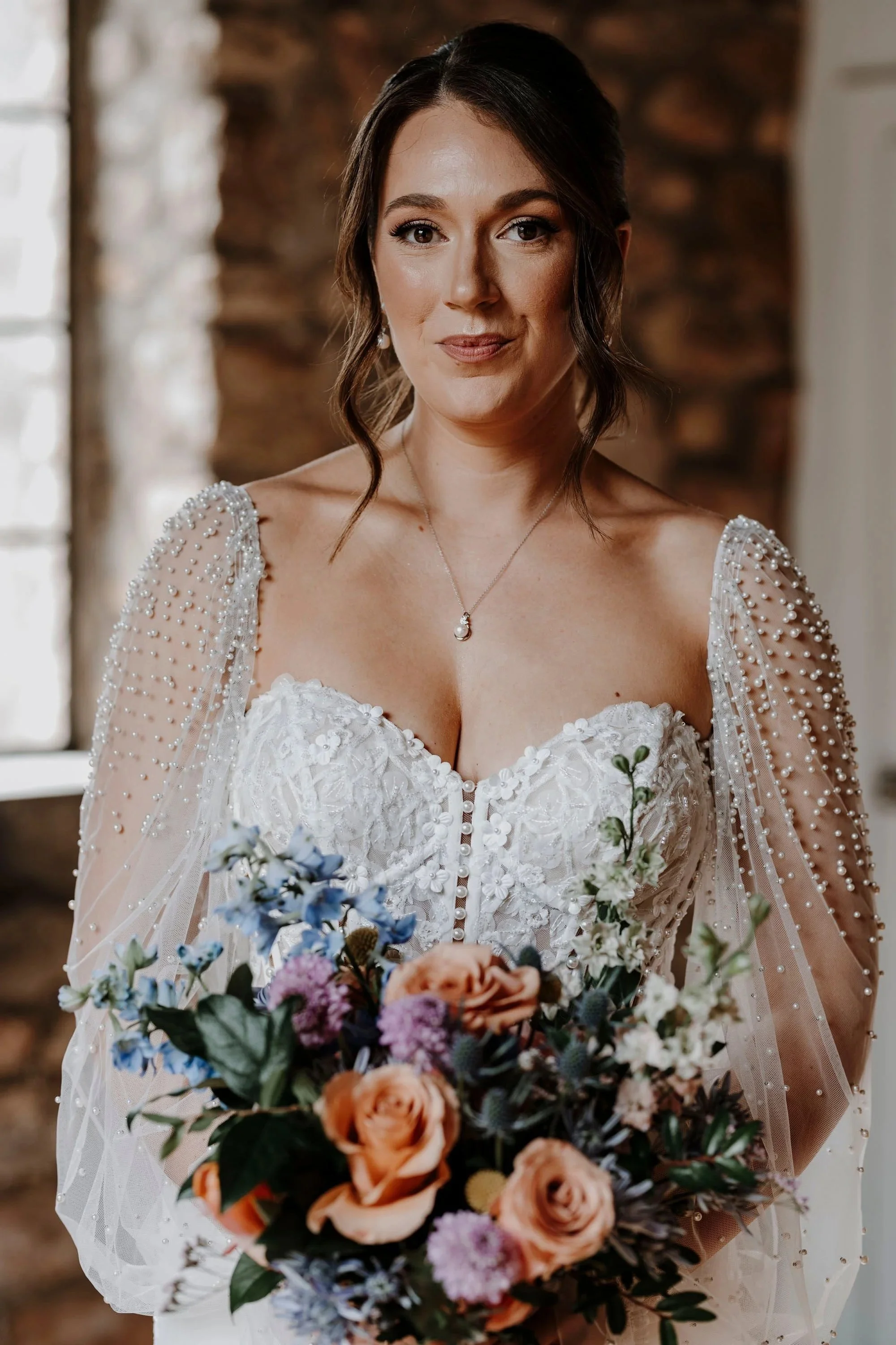 A bride in a white wedding gown with pearl-beaded sheer sleeves, holding a colorful bouquet of flowers, standing in a rustic indoor setting.