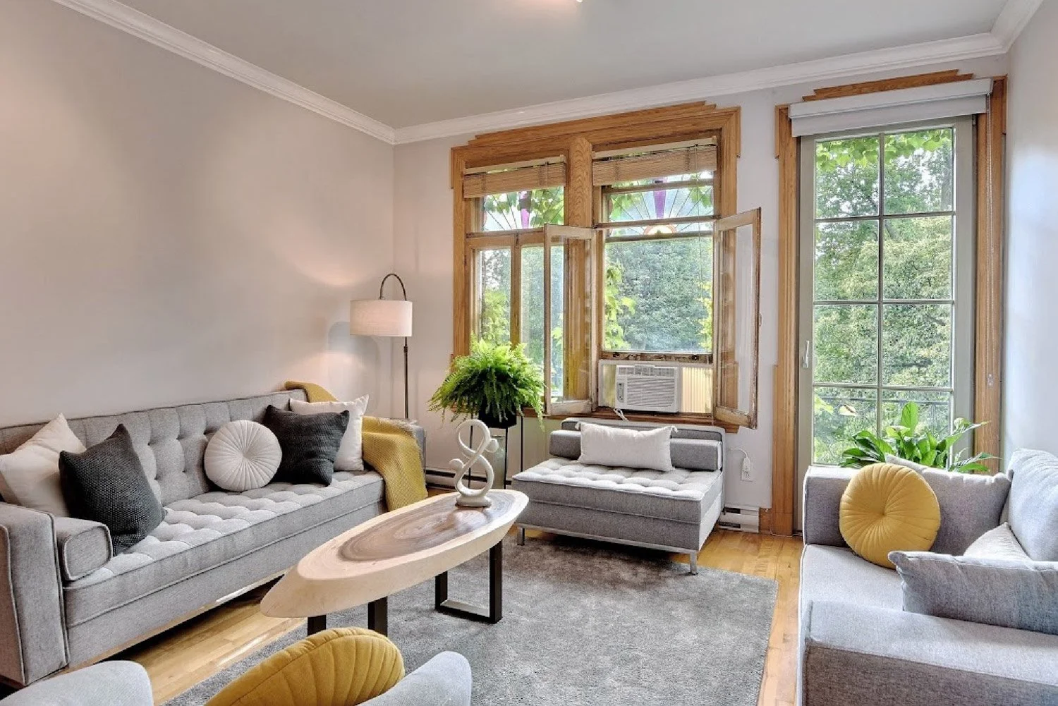 Living room with large windows, gray sofas, decorative pillows, a wooden coffee table, a floor lamp, and green plants.