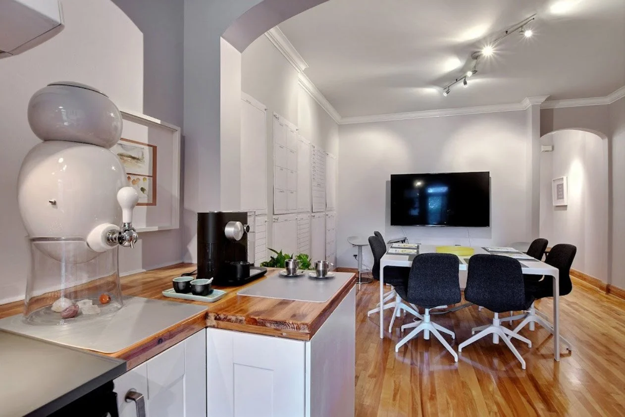 Coffee station with a water dispenser, cups, and a Keurig coffee maker in a modern office room with a large TV and a meeting table.