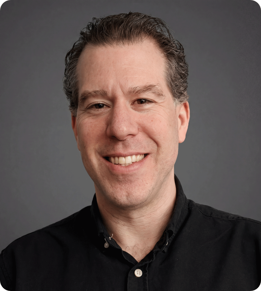 Close-up of a smiling man with dark hair, wearing a black collared shirt, against a gray background.