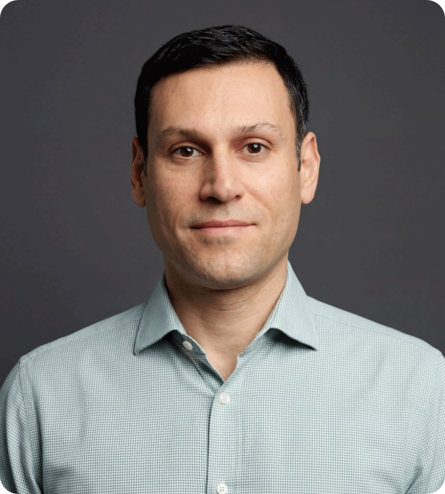 Headshot of a man with short dark hair, wearing a light green dress shirt with a subtle checkered pattern, against a dark grey background.