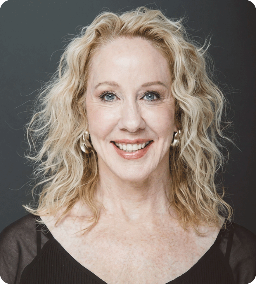 Portrait of a smiling woman with blonde, curly hair wearing a black top with sheer sleeves and pearl earrings against a dark background.